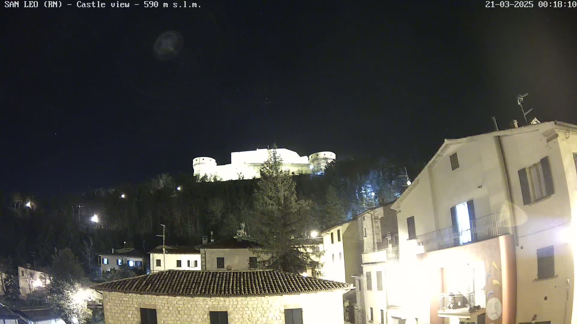 A nighttime view of a hilltop castle illuminated against a dark, clear sky overlooking a small town with buildings lit up.
