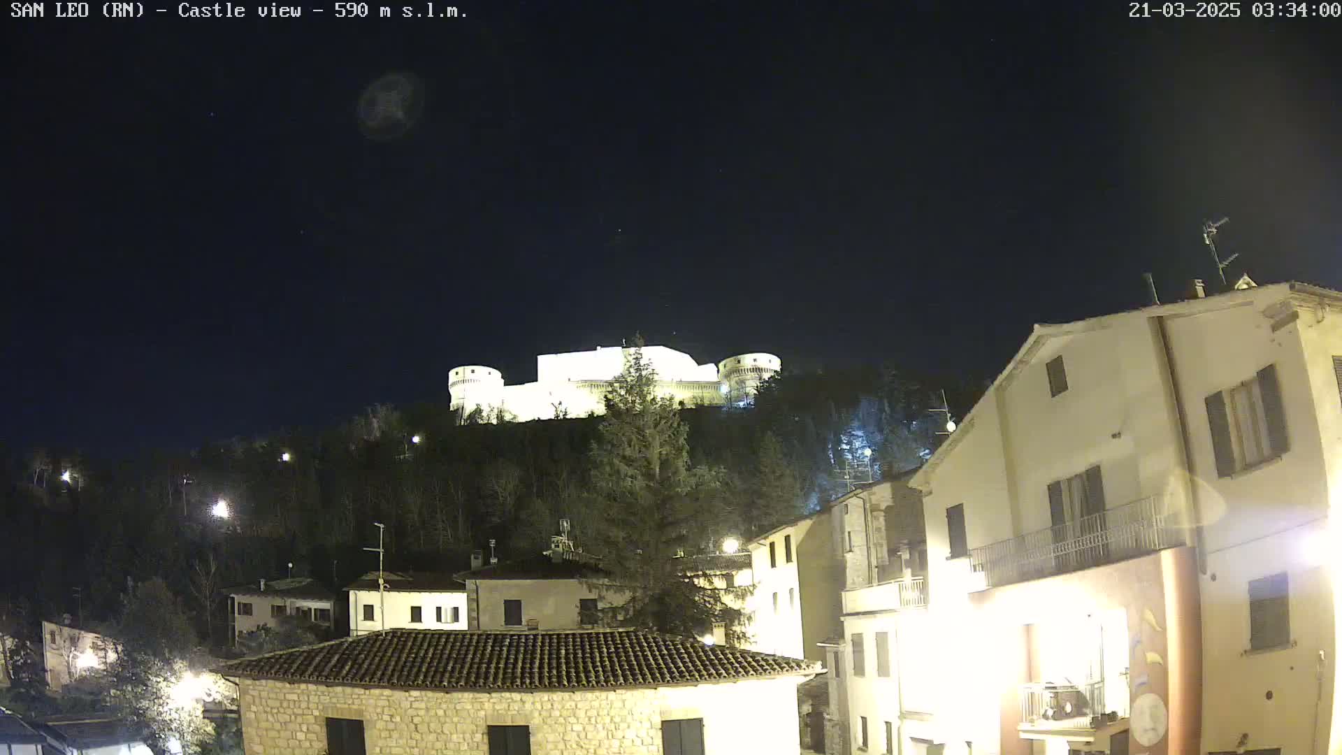 A brightly lit castle sits atop a hill overlooking a town at night under a clear, dark sky.