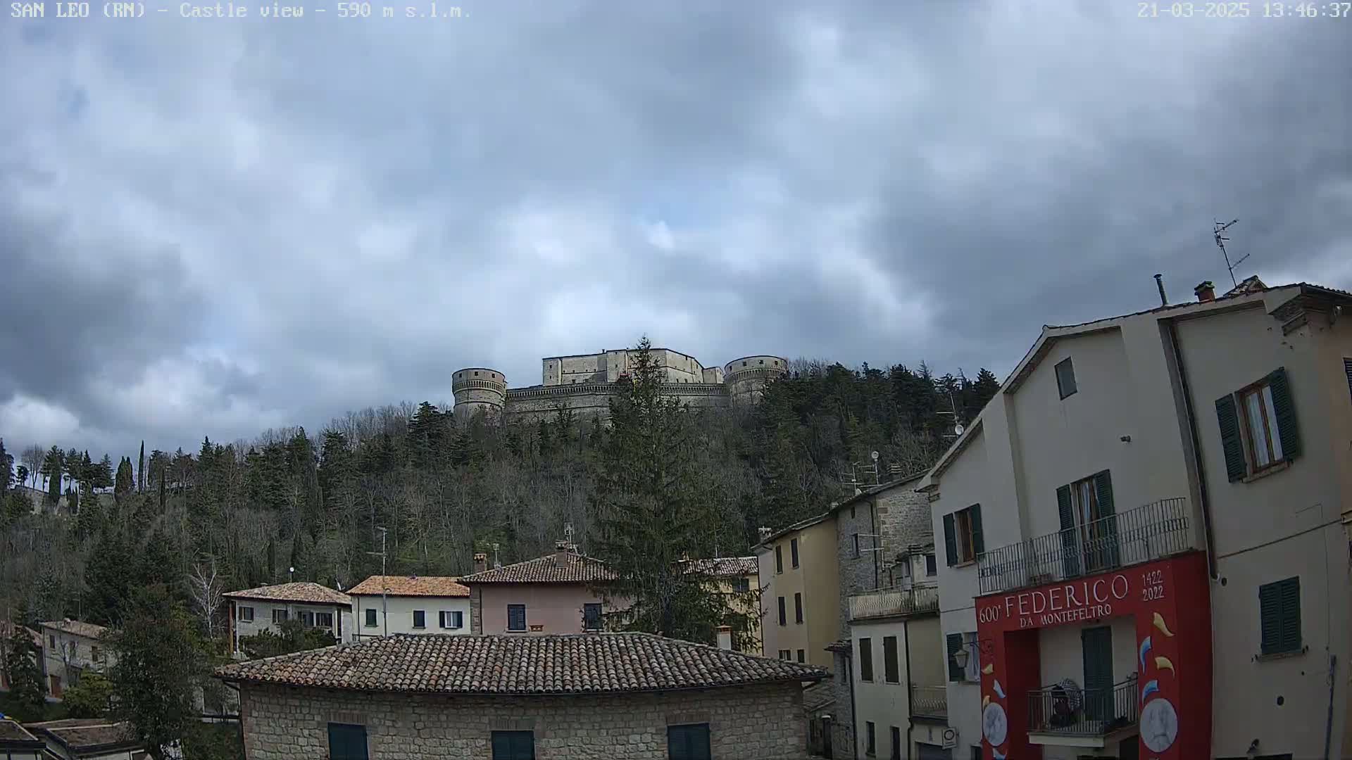 A large stone castle sits atop a tree-covered hill under a mostly cloudy sky, overlooking a town of stone buildings with terracotta roofs.