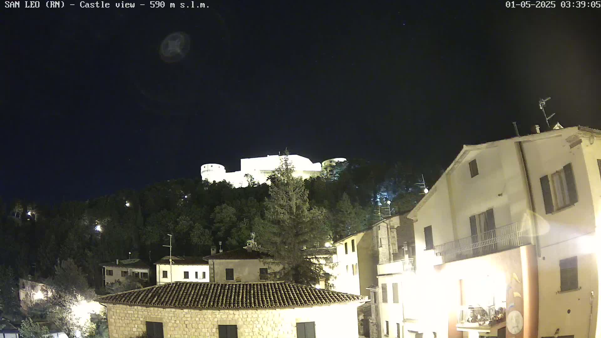 A nighttime view of a hilltop castle illuminated against a dark, clear sky, overlooking a town with buildings lit up below.