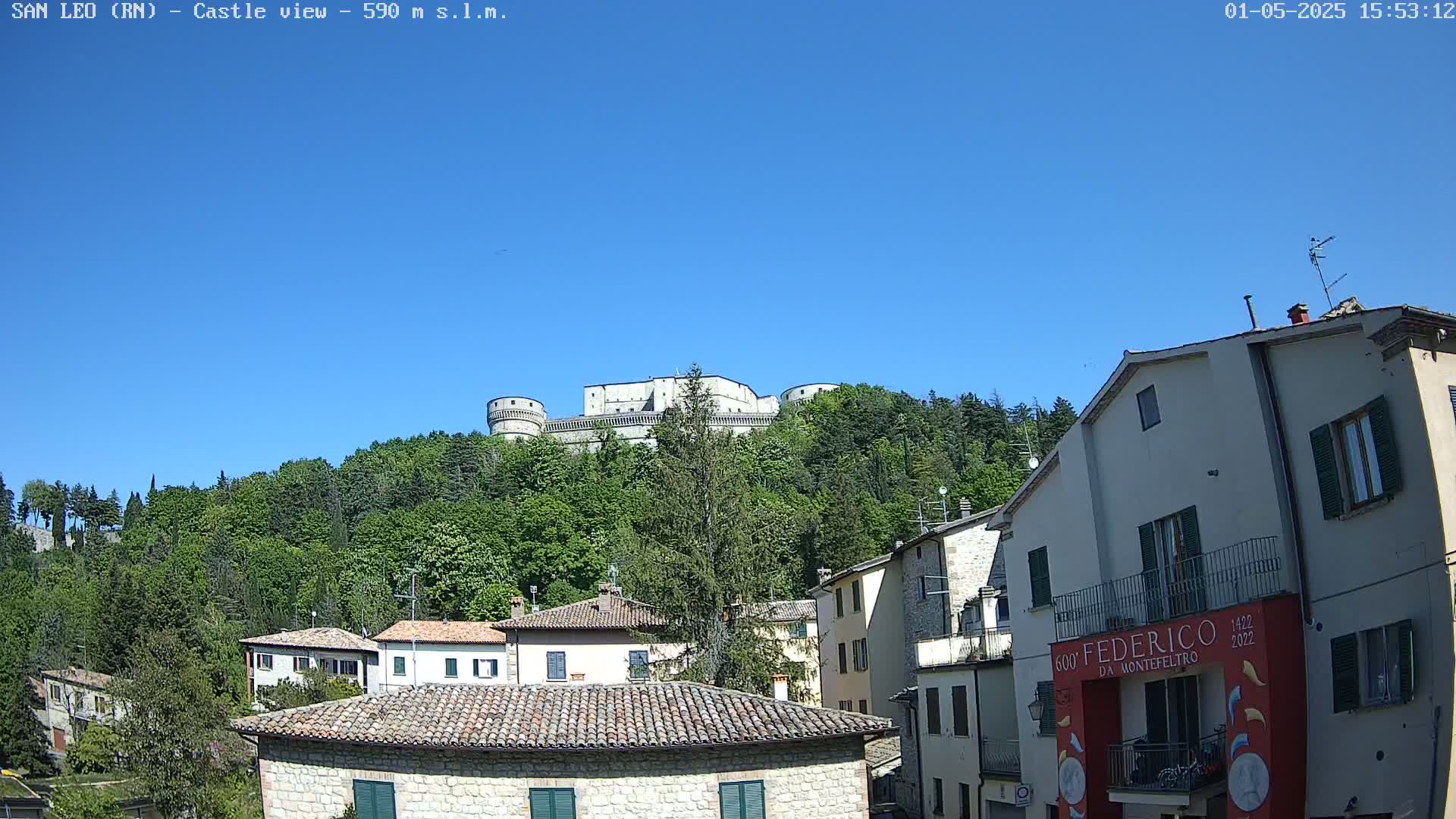 A large stone castle sits atop a hill covered in lush green trees under a clear blue sky, overlooking a village of stone buildings.