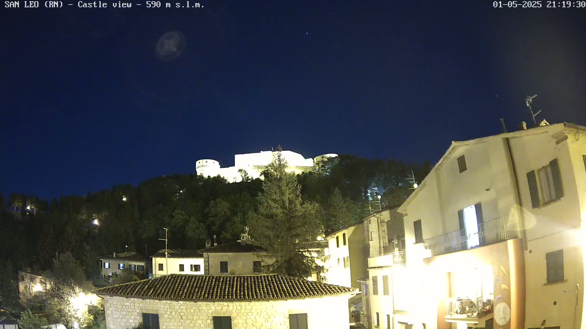 A brightly lit castle sits atop a hill overlooking a village at night under a clear, dark sky.