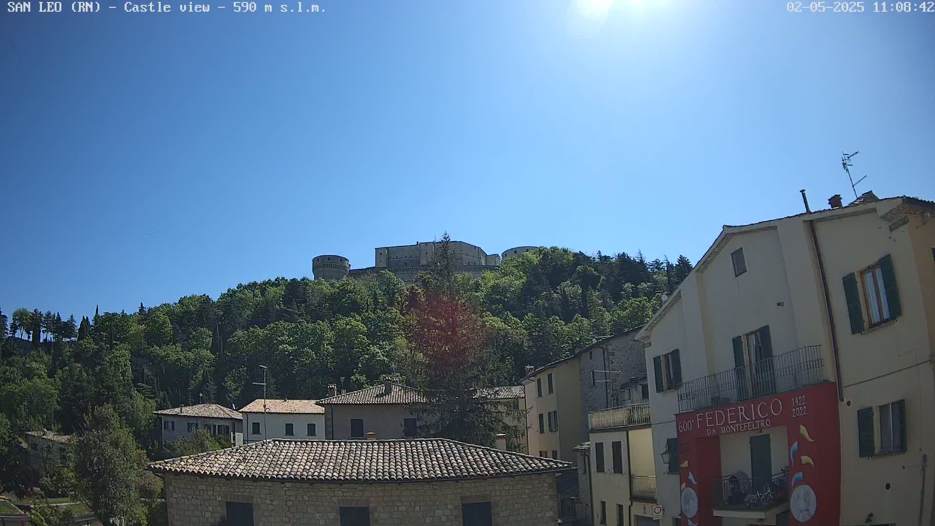 A stone castle sits atop a hill covered in lush green trees under a clear blue sky, with a village of stone buildings nestled at its base.