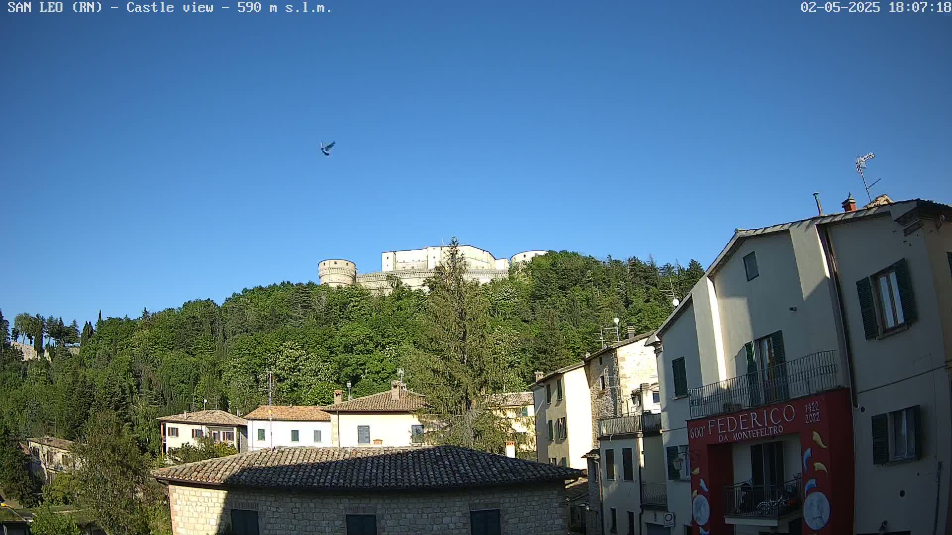 A stone castle sits atop a verdant hill overlooking a town of low stone buildings under a clear blue sky.