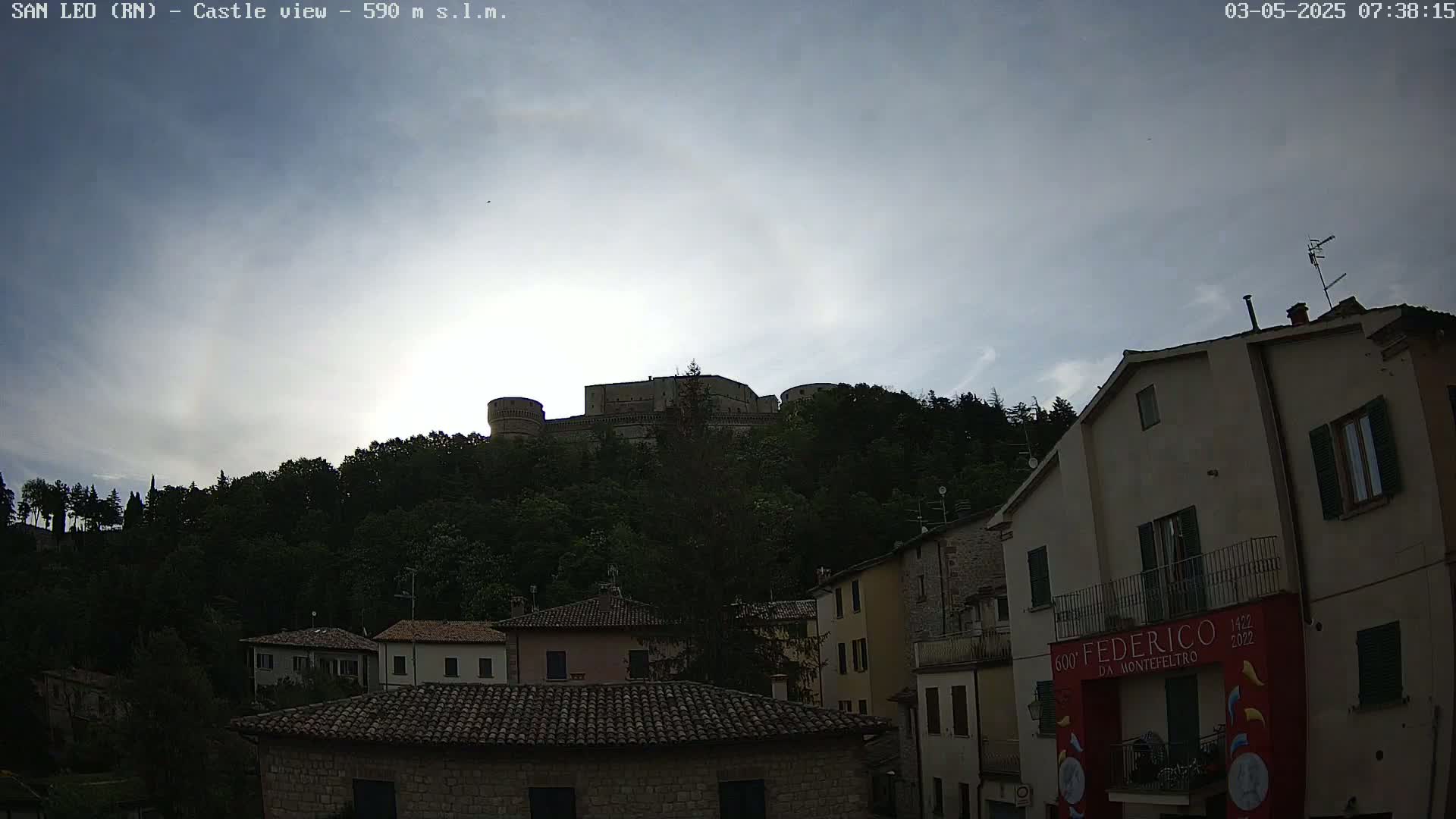 A stone castle sits atop a hill covered in lush green trees under a mostly cloudy sky.