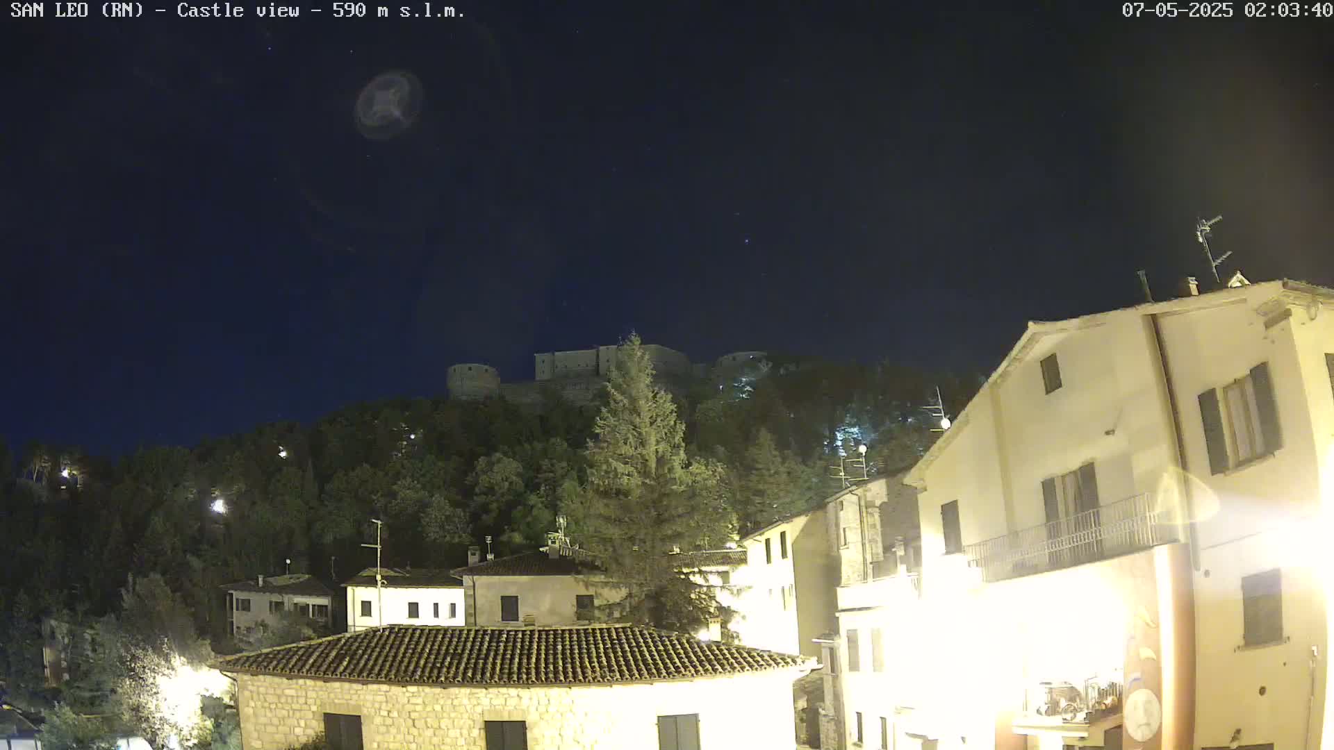 A nighttime view of a hilltop castle overlooking a town with illuminated buildings under a clear, dark sky.