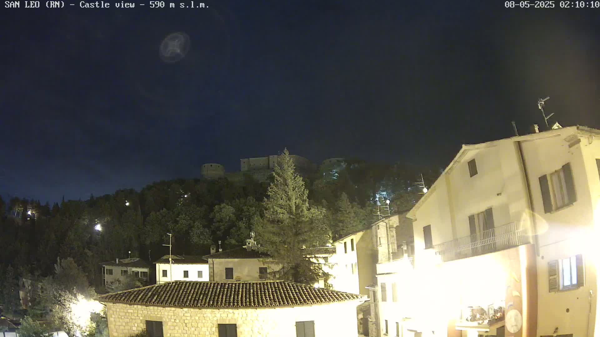 A nighttime view of a hilltop castle overlooking a town with buildings illuminated under a clear, dark sky.