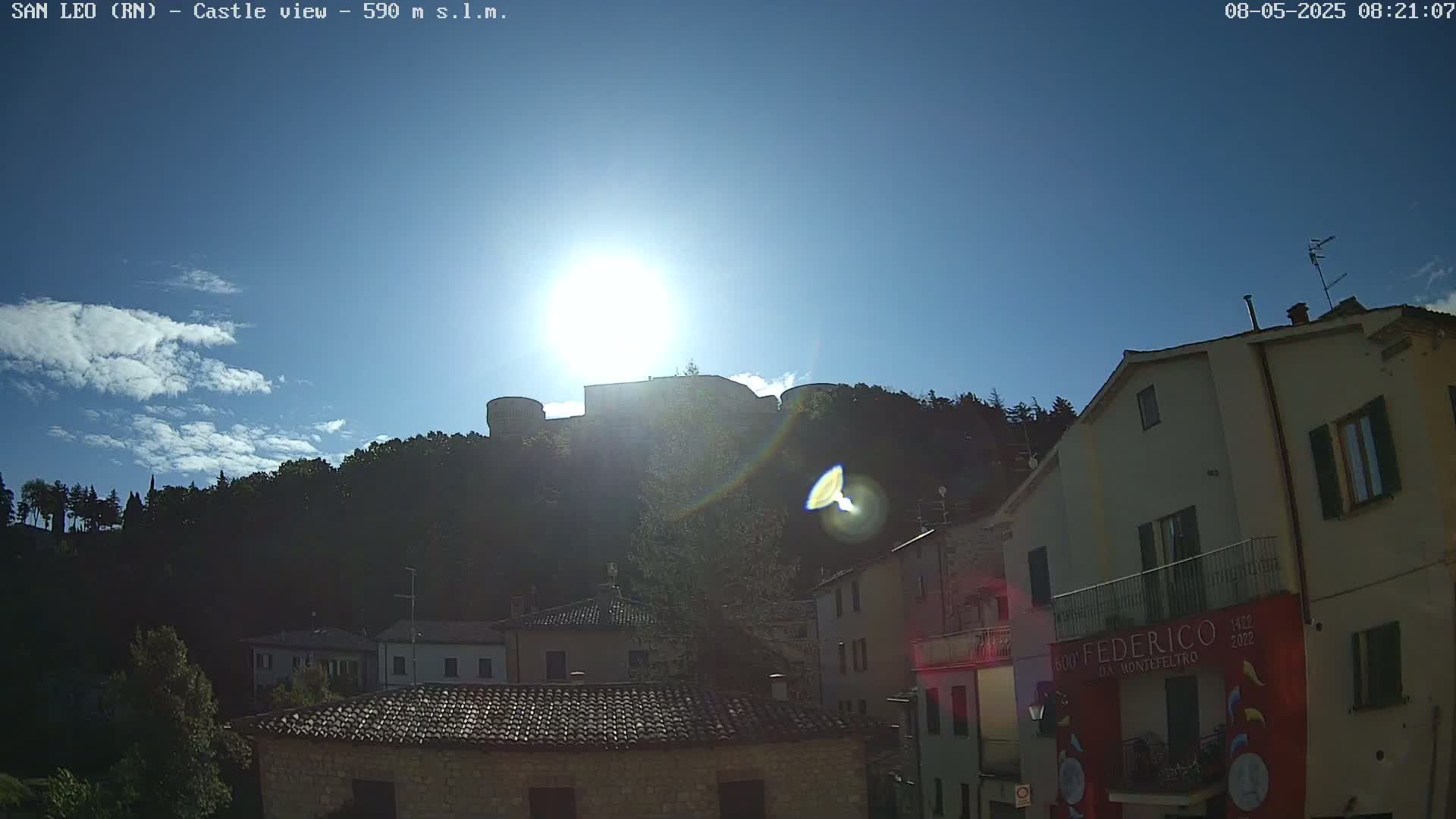 A hilltop castle is silhouetted against a bright, sunny sky with a few scattered clouds, overlooking a town of low buildings with terracotta roofs.