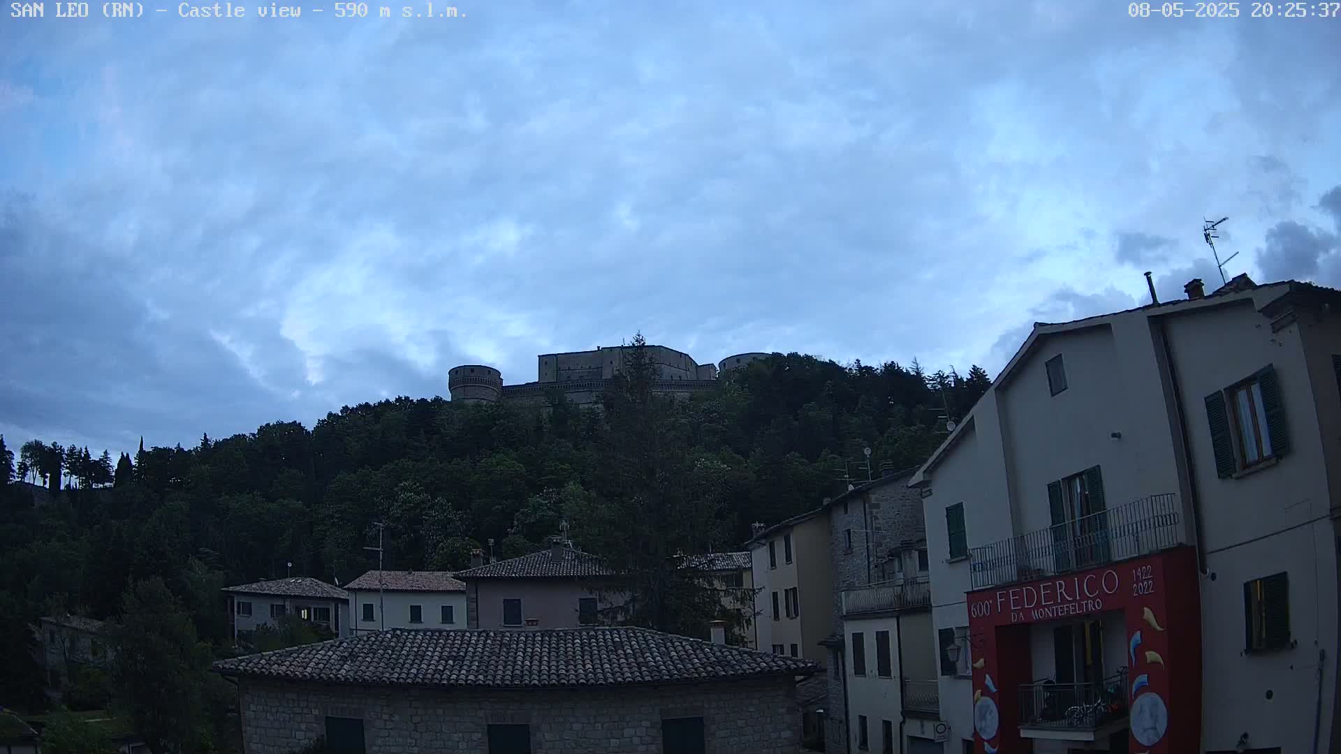 A stone castle sits atop a hill covered in trees under a mostly cloudy, twilight sky, overlooking a village of stone buildings.