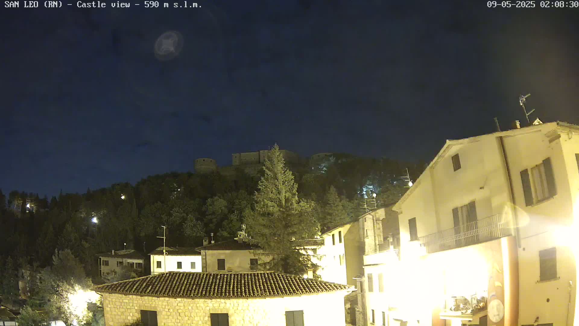 A nighttime view of a hilltop castle overlooking a town with buildings illuminated under a dark, clear sky.