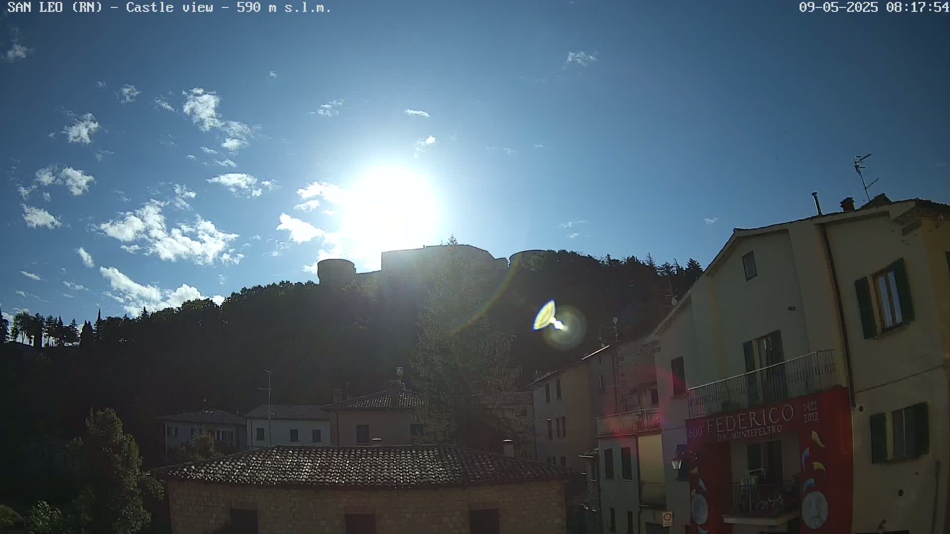 A hilltop castle is silhouetted against a bright, sunny sky with scattered clouds, overlooking a village of buildings with terracotta roofs.