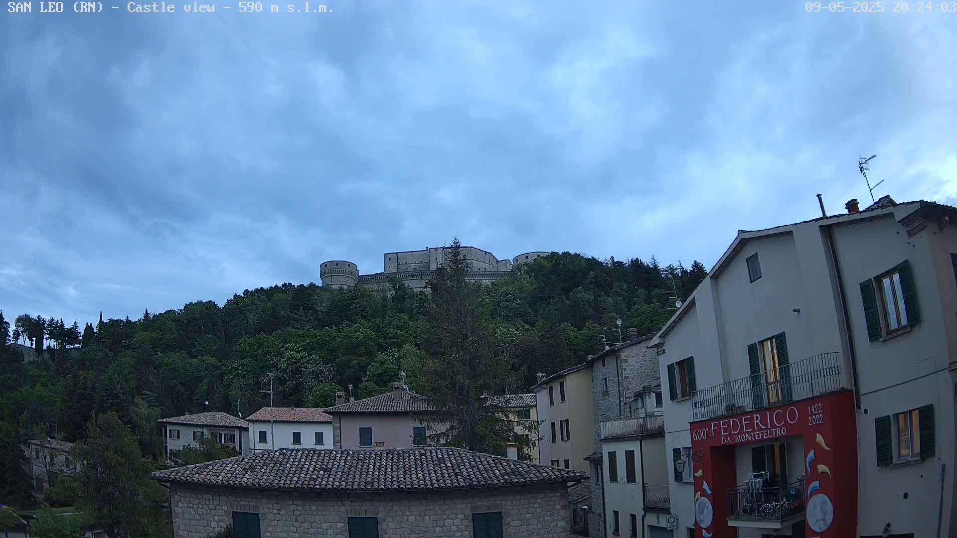 A stone castle sits atop a tree-covered hill under a mostly cloudy, twilight sky, overlooking a town of stone buildings.