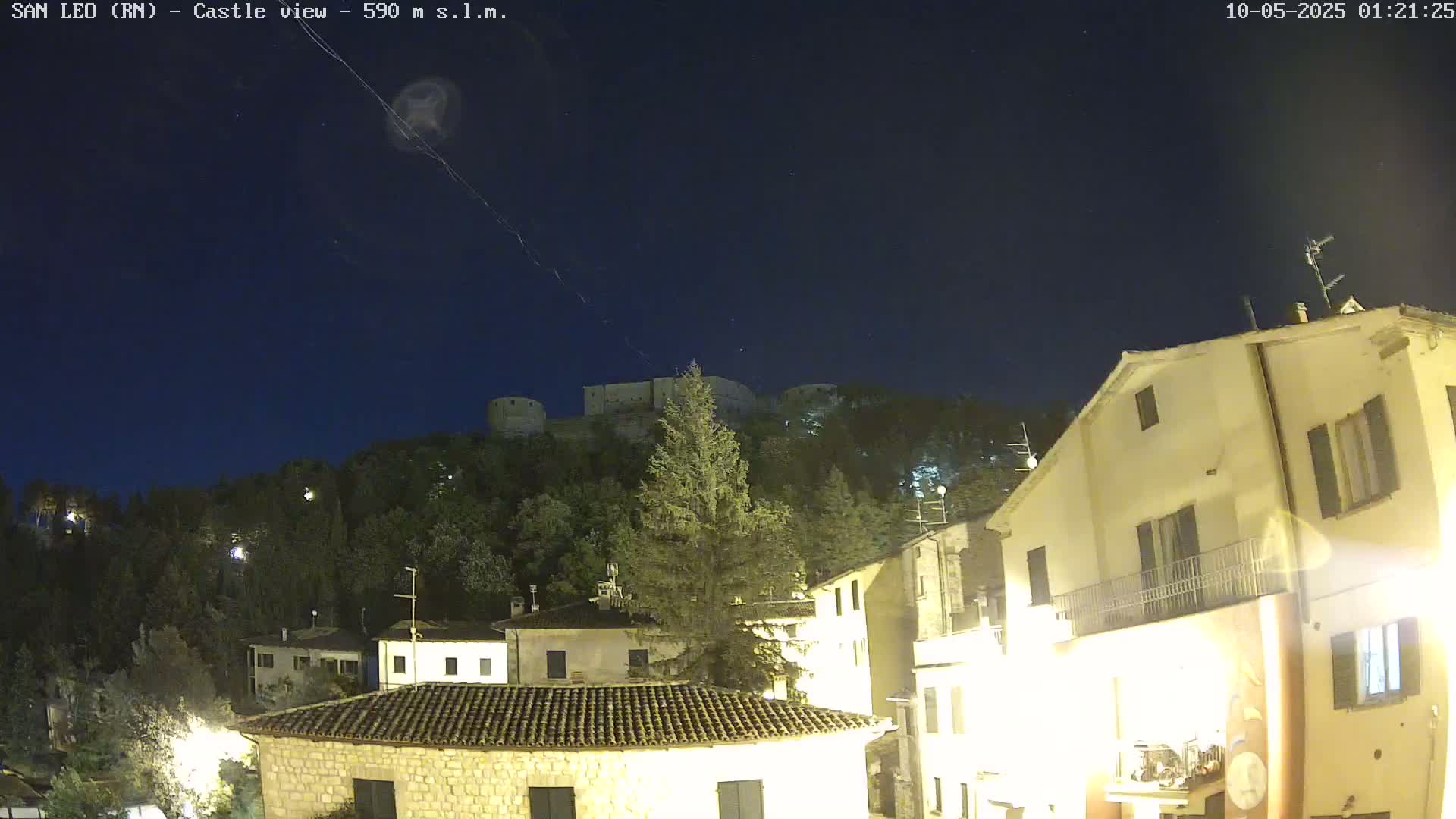 A nighttime view of a hilltop castle overlooking a village of illuminated buildings under a clear, starry sky.