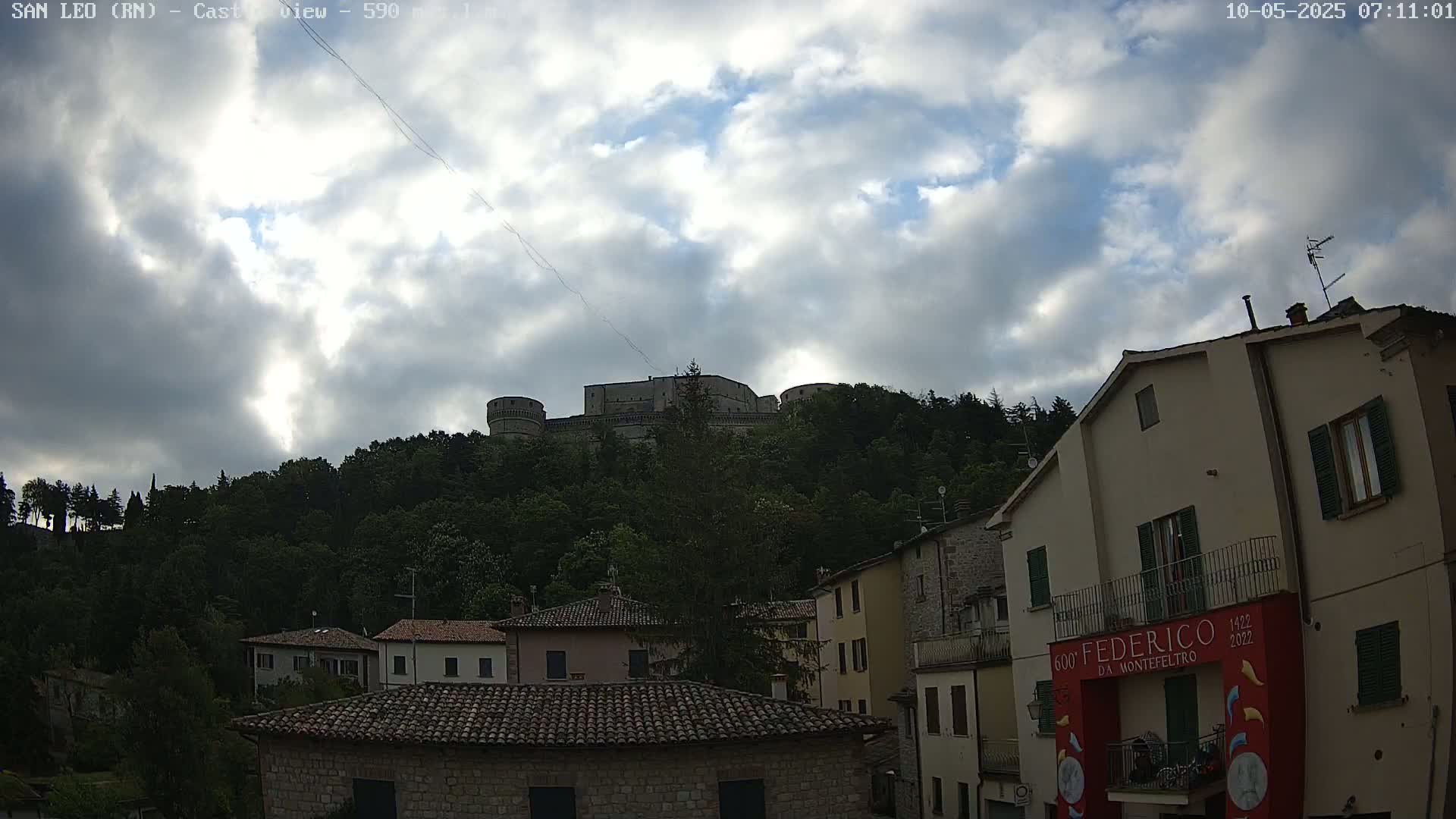 A stone castle sits atop a hill covered in lush green trees under a partly cloudy sky, overlooking a town of closely-packed stone buildings.