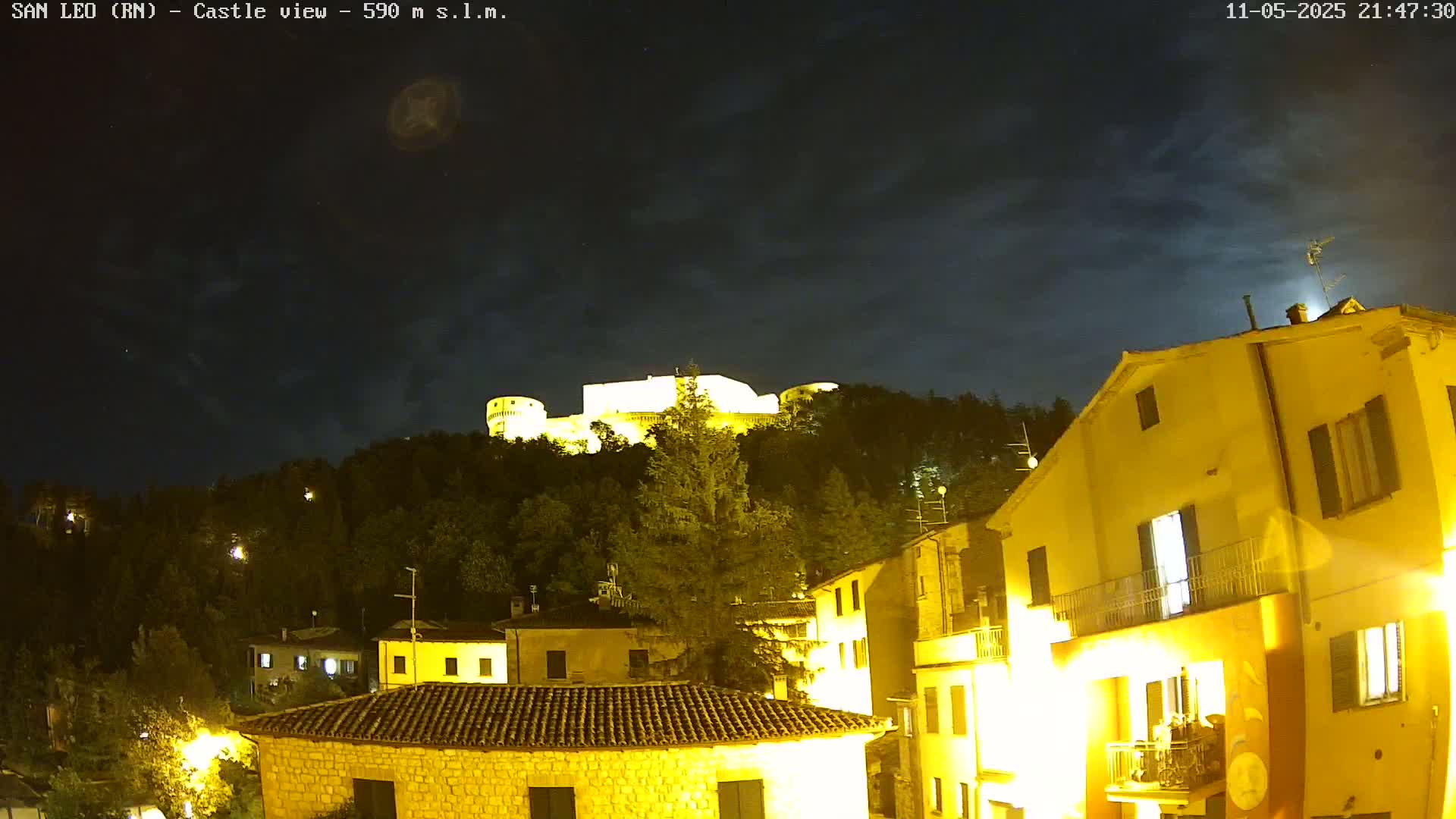 A hilltop castle is illuminated at night under a partly cloudy sky, overlooking a town with brightly lit buildings.
