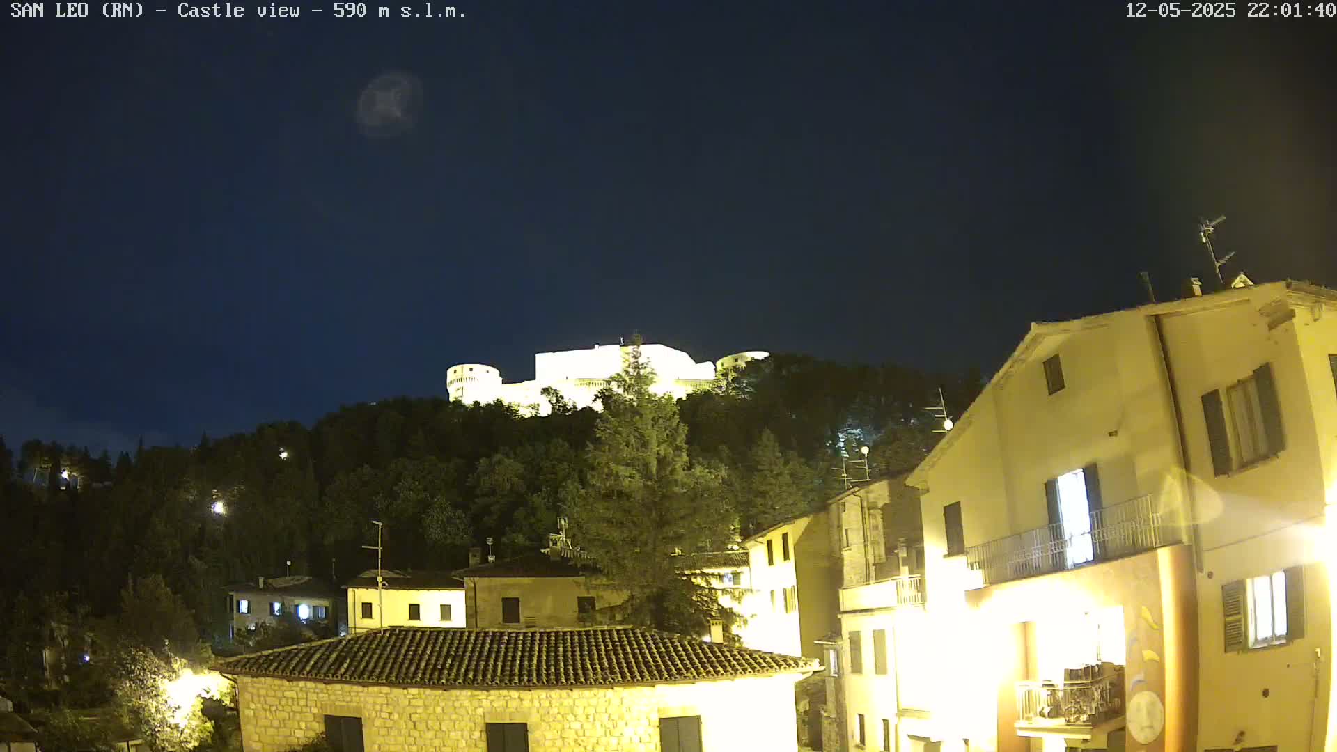 A nighttime view of a hilltop castle illuminated against a clear, dark sky, overlooking a village of lit buildings.