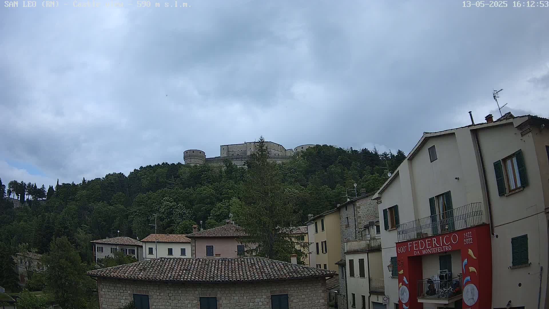 A stone fortress sits atop a hill covered in lush green trees under a cloudy sky, overlooking a village of stone buildings with red-tiled roofs.