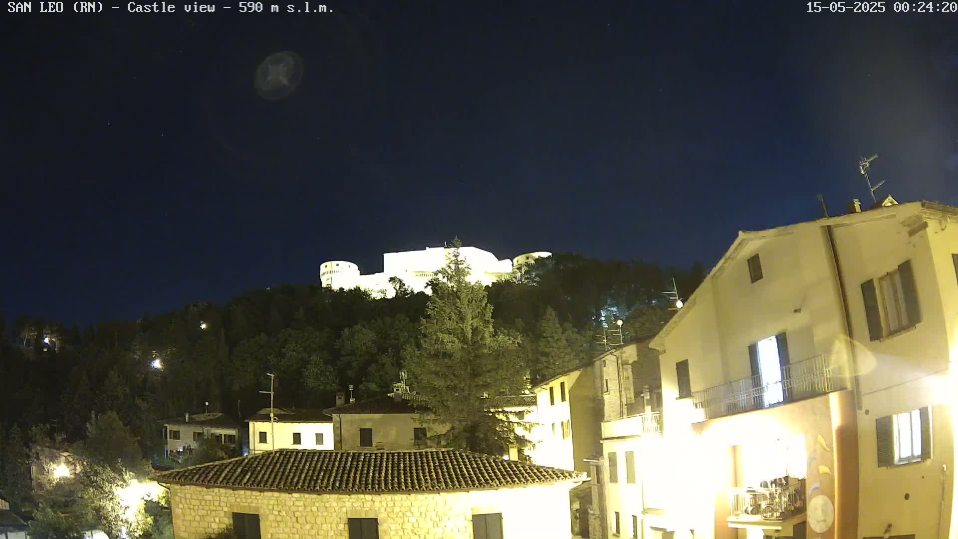 A nighttime view of a hilltop castle illuminated against a dark, clear sky, with the lights of a village visible in the foreground.