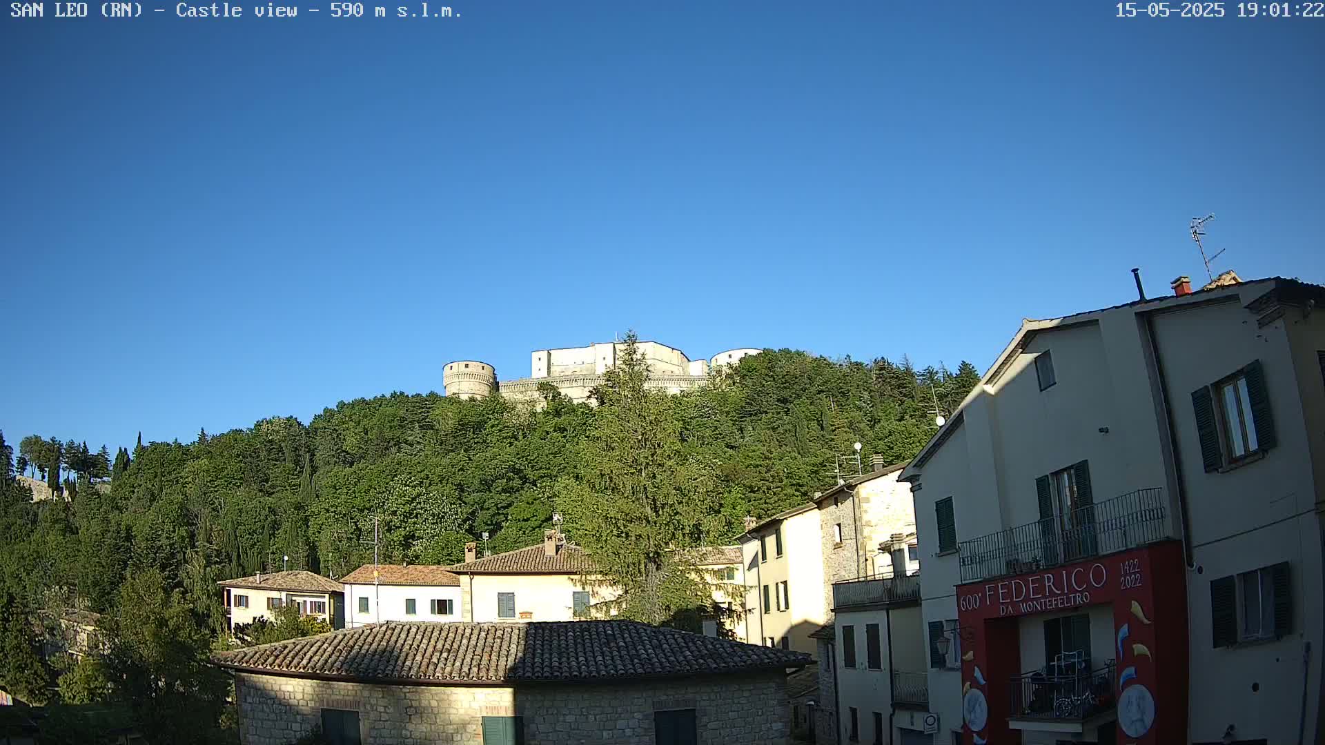 A stone castle sits atop a hill covered in green trees under a clear blue sky, overlooking a village of stone buildings.