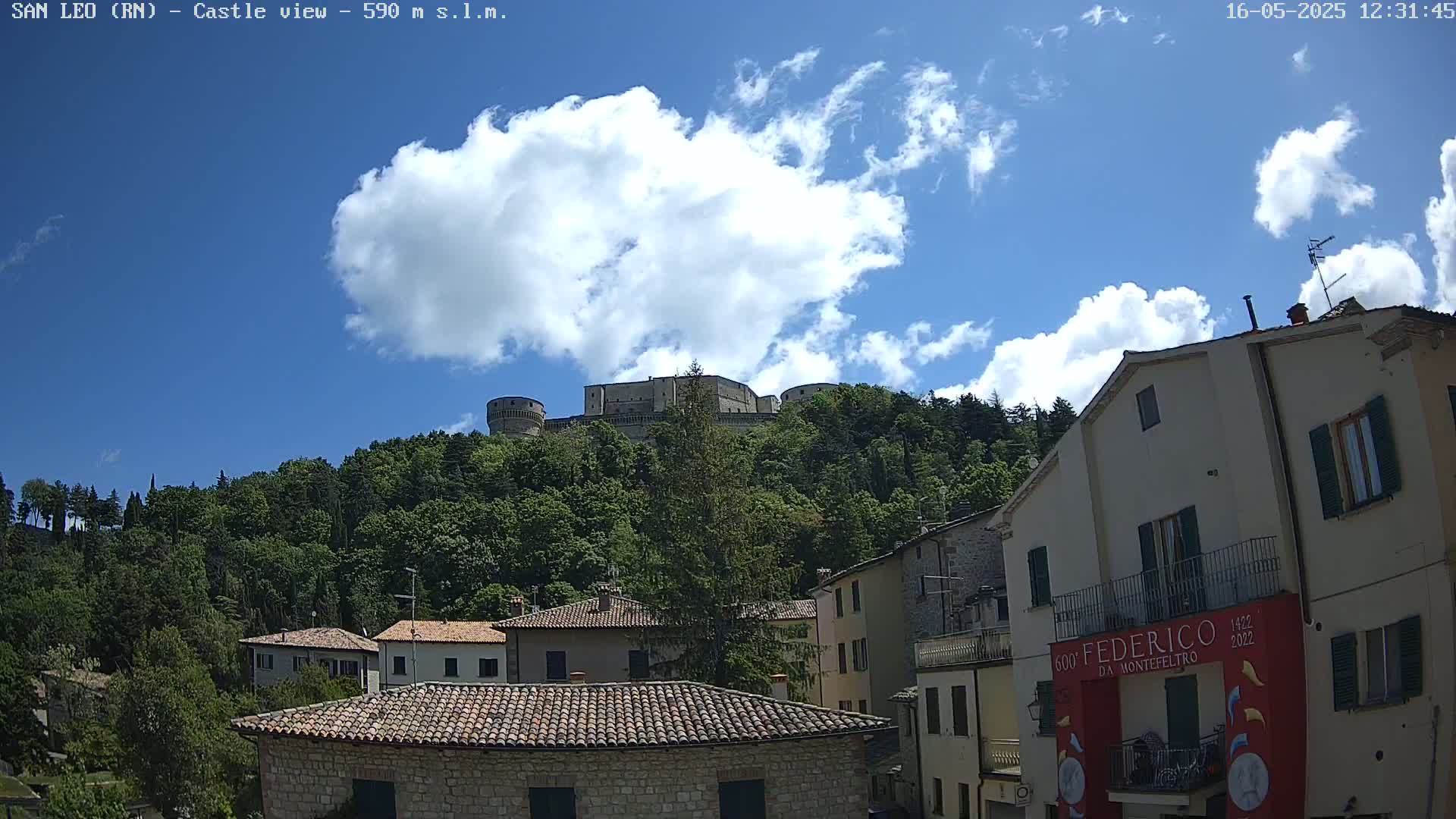 A stone castle sits atop a tree-covered hill under a partly cloudy blue sky, overlooking a town of stone buildings with red-tiled roofs.