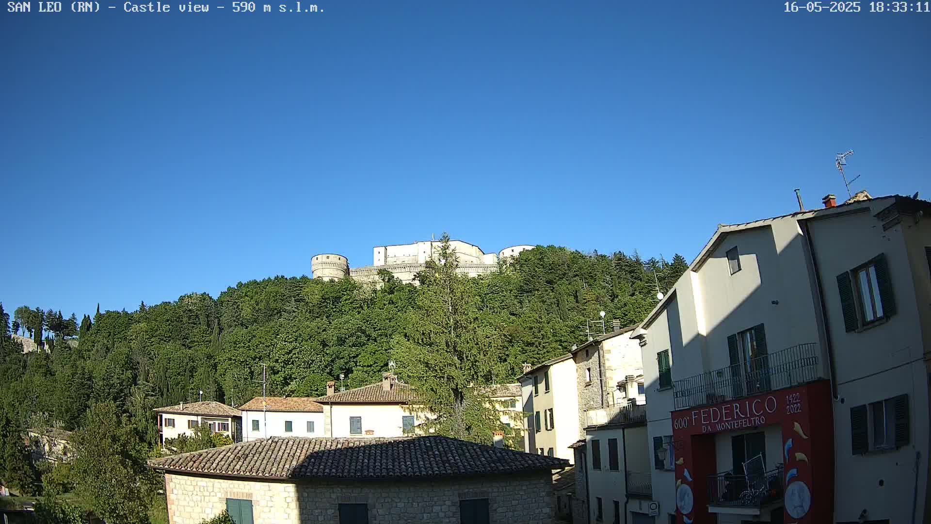 A stone castle sits atop a hill covered in lush green trees, overlooking a town of low stone buildings under a clear blue sky.