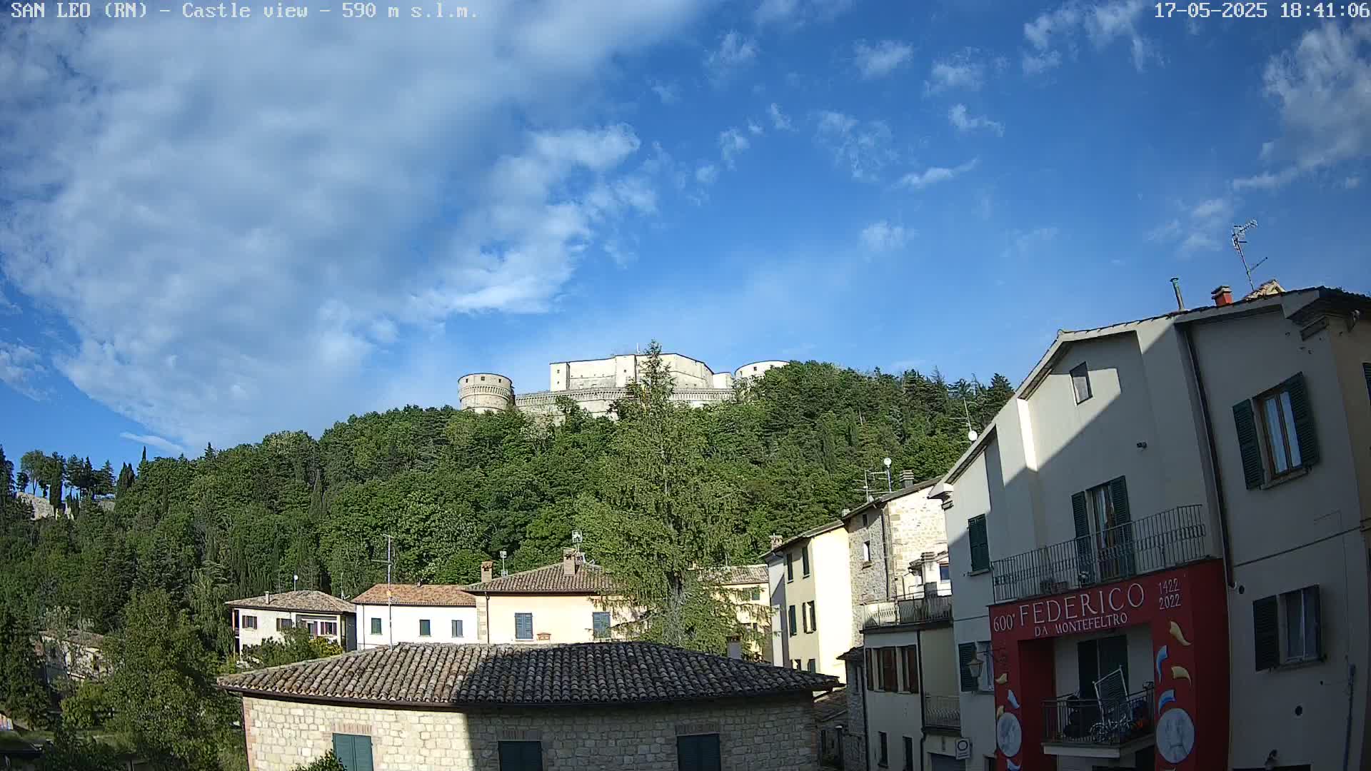 A stone castle sits atop a tree-covered hill overlooking a town of low stone buildings under a mostly sunny sky with scattered clouds.
