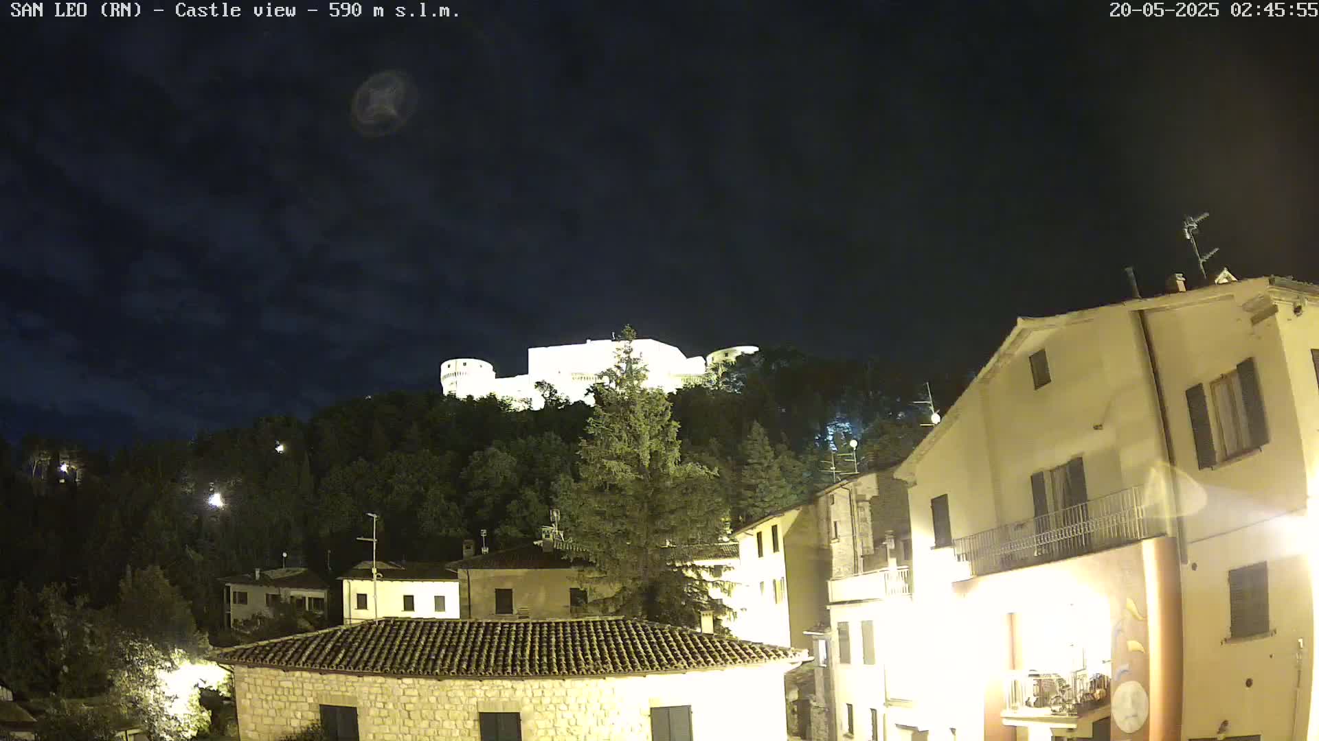 A nighttime view of a hilltop castle illuminated against a dark, cloudy sky, overlooking a village with several buildings lit up.