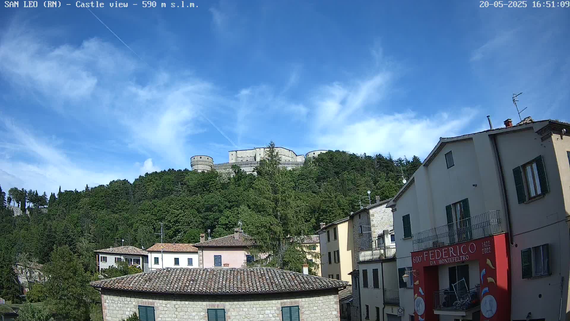 A stone castle sits atop a verdant hill under a mostly clear blue sky with wispy clouds, overlooking a village of stone buildings.