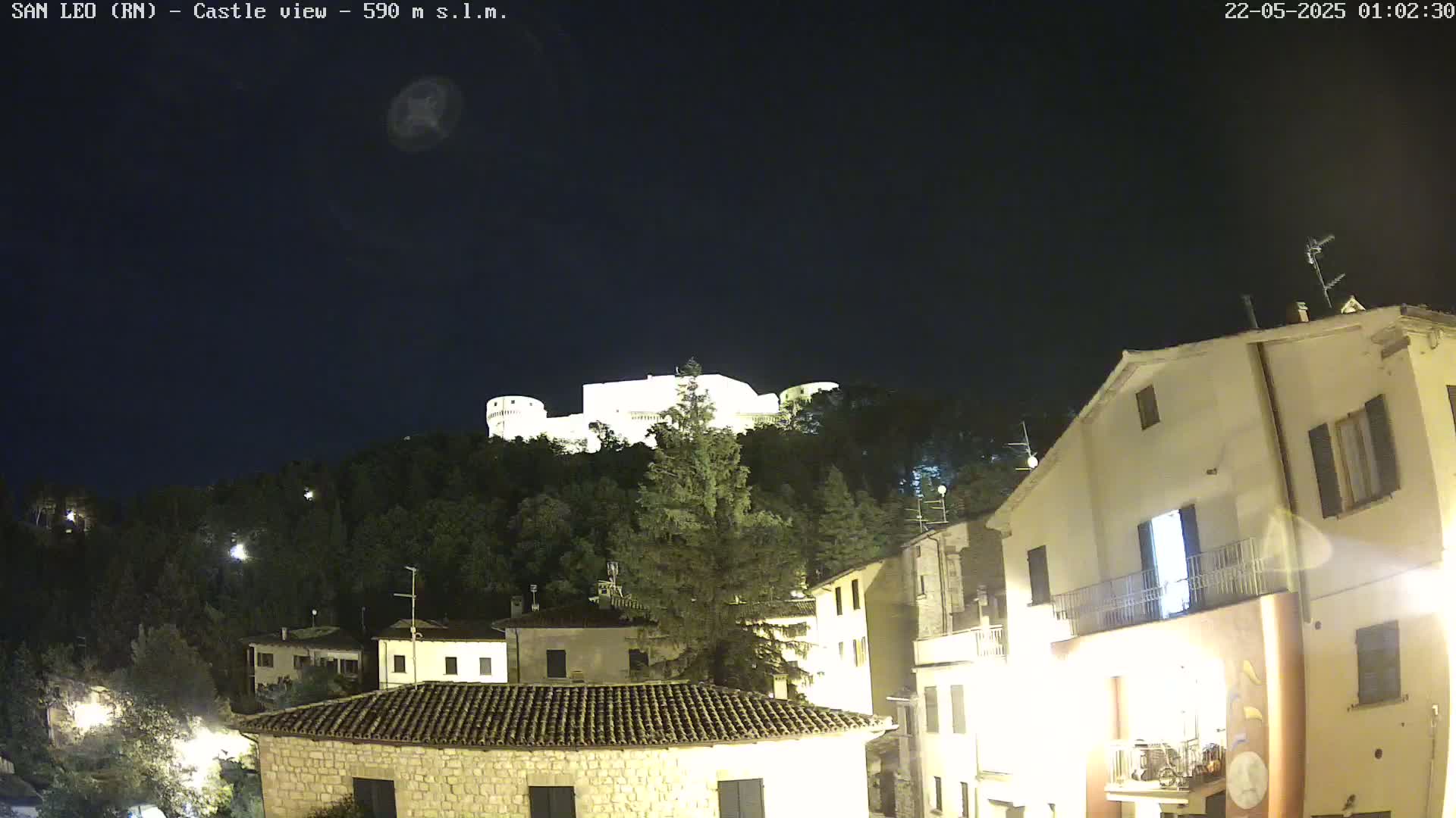 A nighttime view of a hilltop castle illuminated against a dark sky, overlooking a village with buildings lit up below.