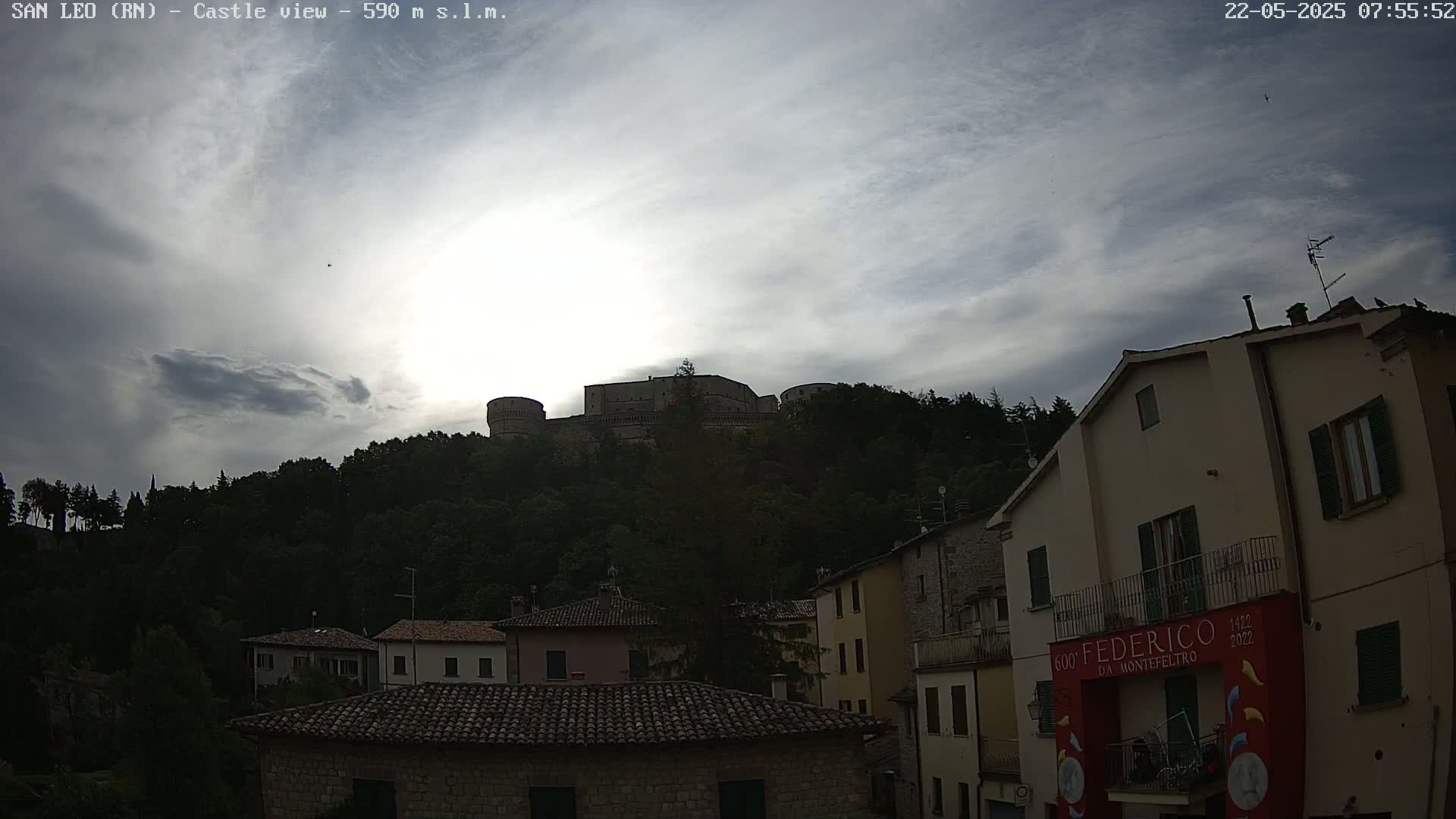 A stone castle sits atop a hill covered in dark green trees under a partly cloudy sky.