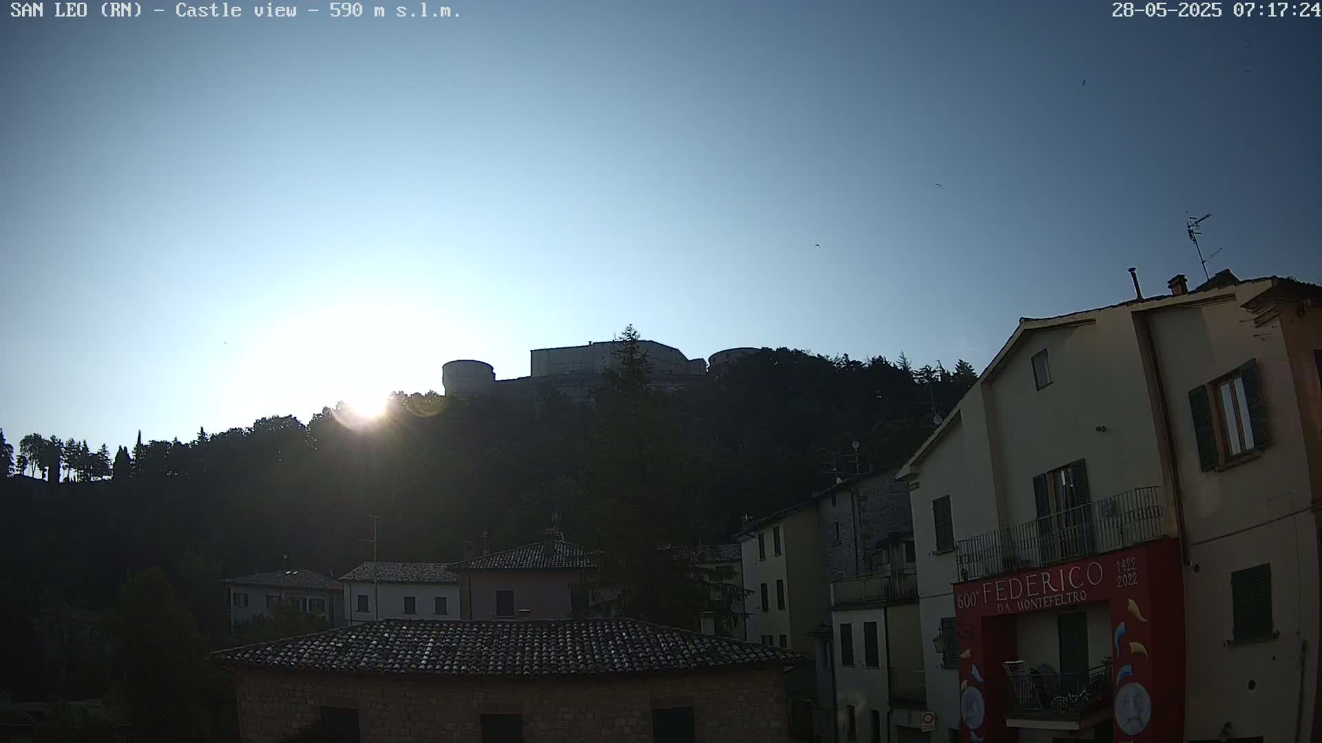 A hilltop castle is silhouetted against a clear, bright sunrise over a town of low buildings.