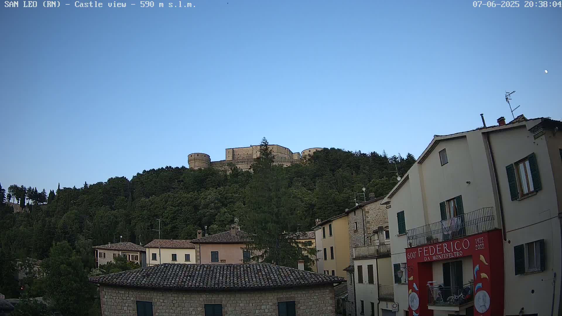 A stone castle sits atop a hill covered in lush green trees under a clear blue twilight sky, overlooking a town with buildings of similar stone construction.