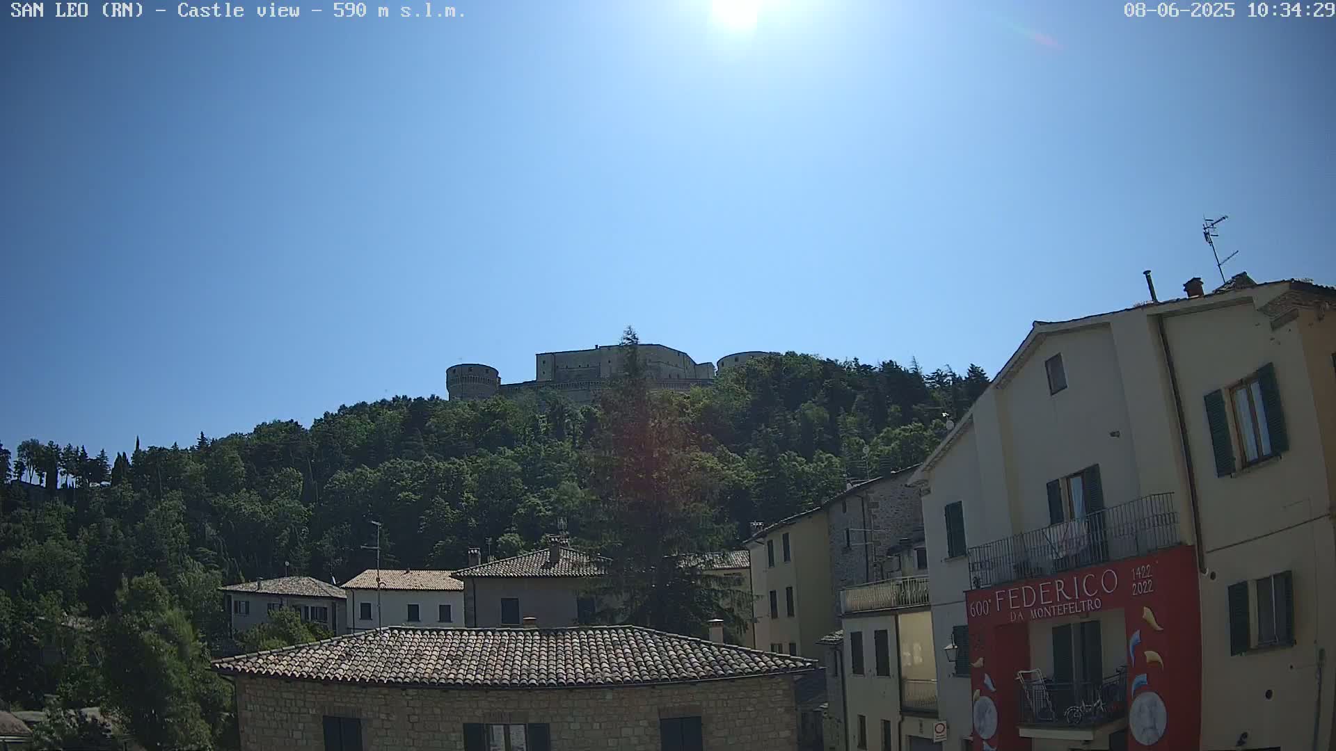 A stone castle sits atop a tree-covered hill under a bright, sunny sky, overlooking a village of stone buildings with terracotta roofs.