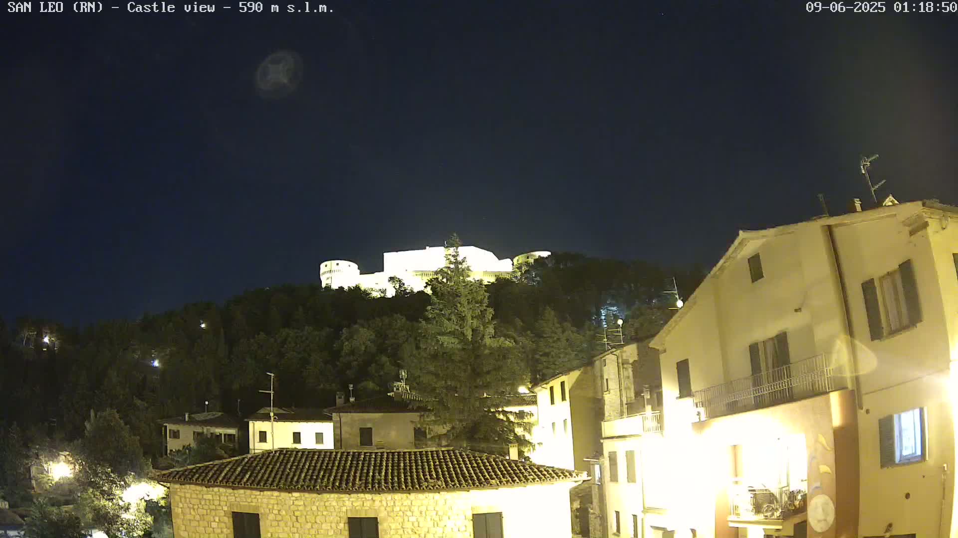 A nighttime view of a hilltop castle illuminated against a dark, clear sky, overlooking a town with buildings lit up below.