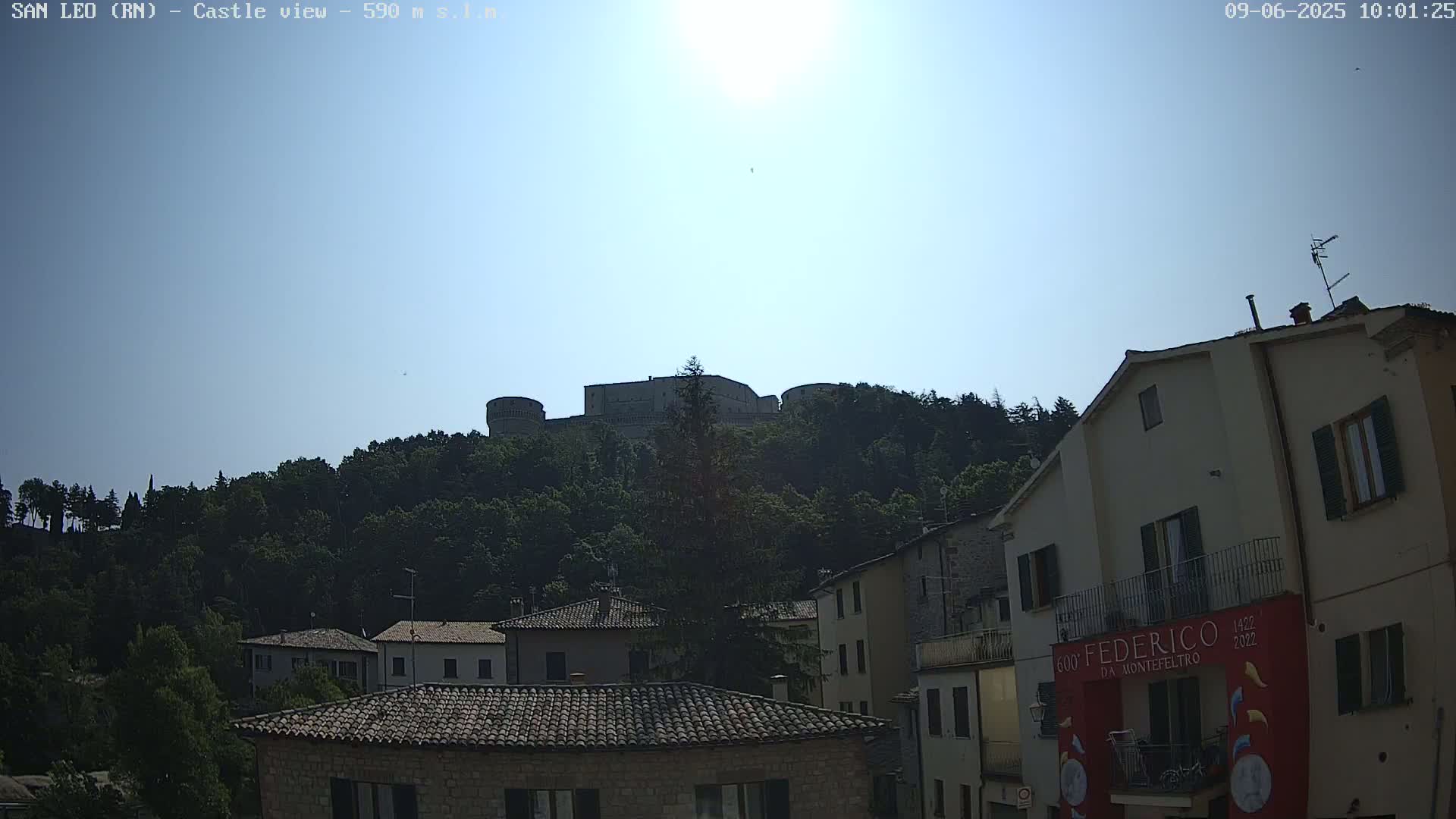 A stone castle sits atop a tree-covered hill under a bright, sunny sky, overlooking a town of low buildings with terracotta roofs.