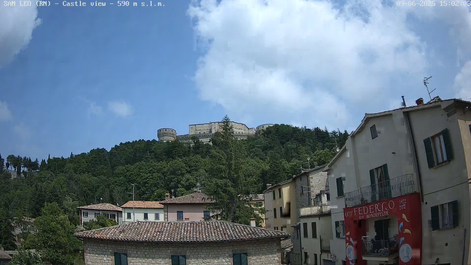 A large stone castle sits atop a hill covered in lush green trees, overlooking a town of low stone buildings under a partly cloudy sky.