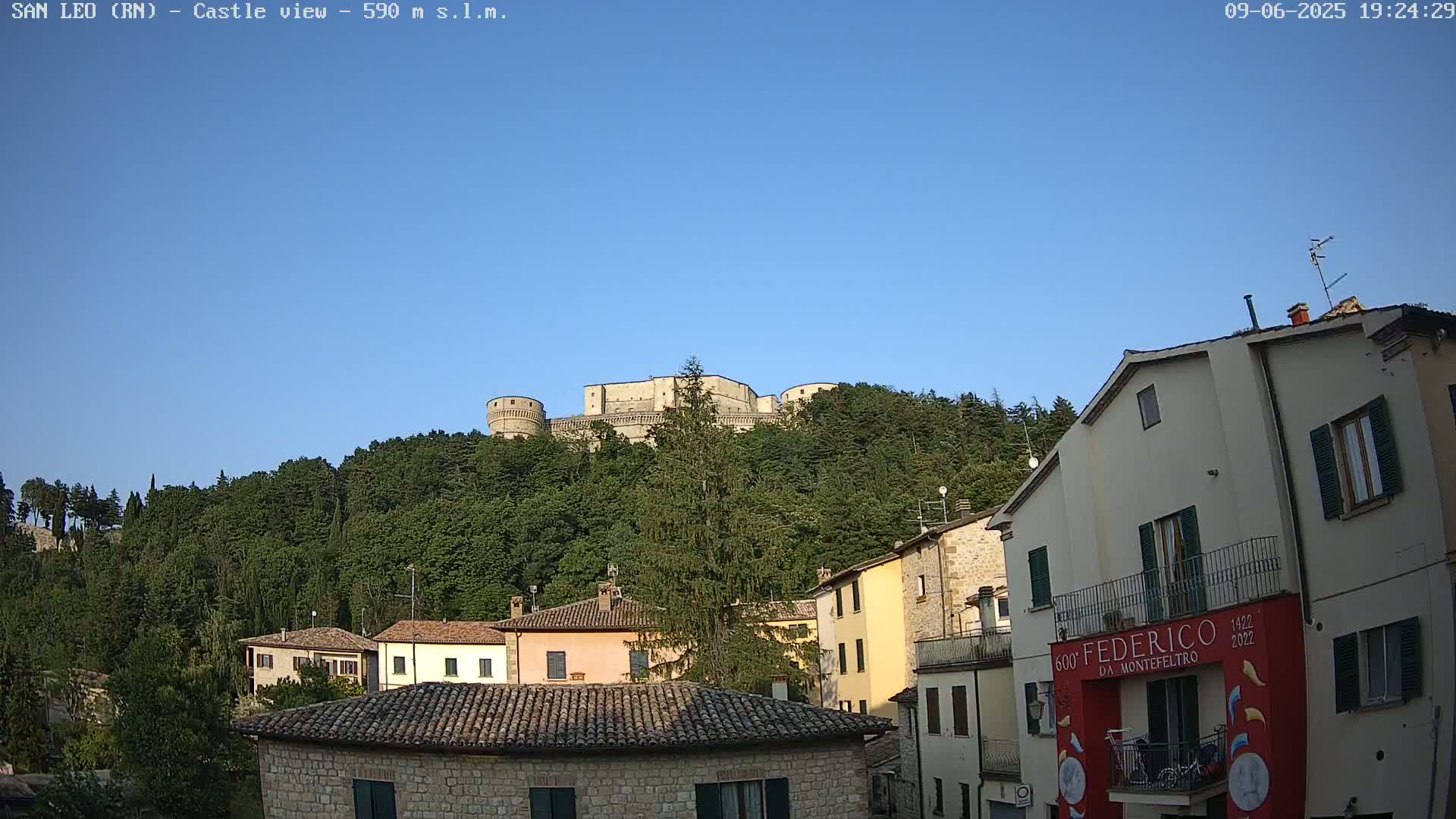 A stone castle sits atop a tree-covered hill overlooking a town of stone buildings under a clear blue sky.