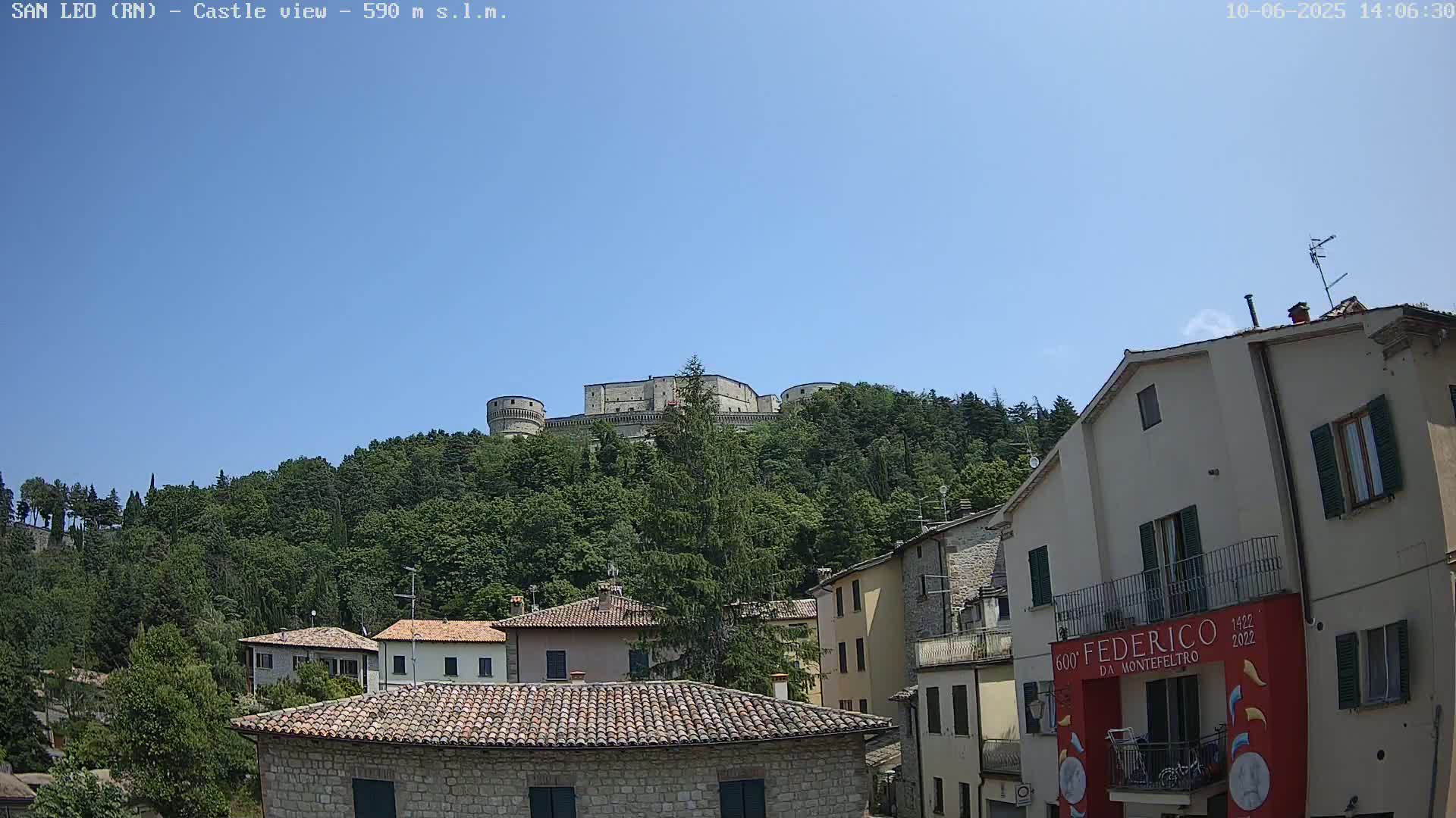 A stone castle sits atop a tree-covered hill overlooking a town of stone buildings under a clear blue sky.