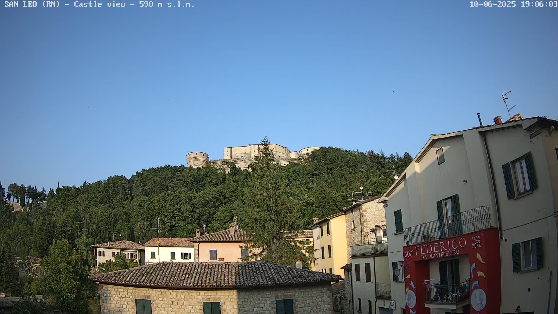 A stone castle sits atop a hill covered in lush green trees under a clear blue sky, overlooking a town with terracotta-roofed buildings.