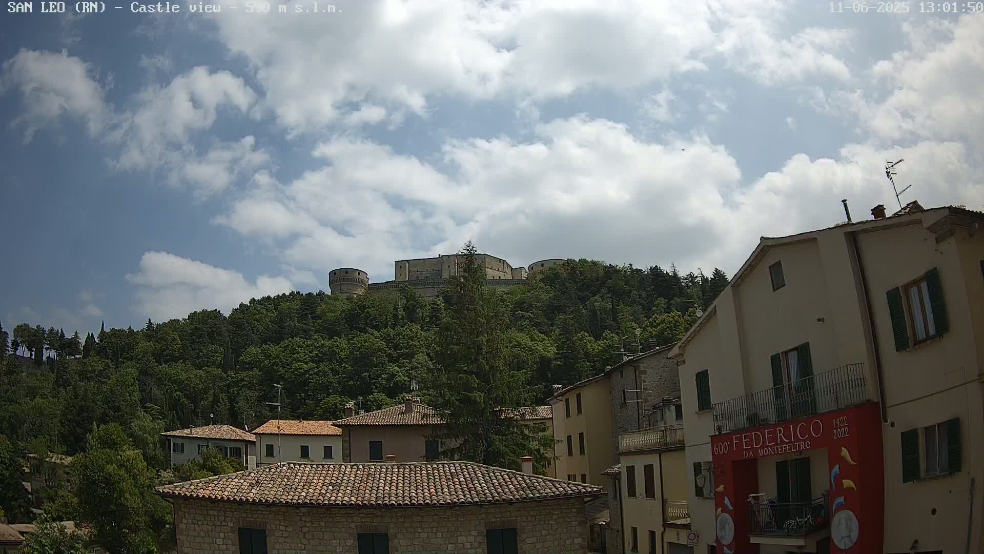 A stone castle sits atop a tree-covered hill under a partly cloudy sky, overlooking a town of stone buildings with terracotta roofs.