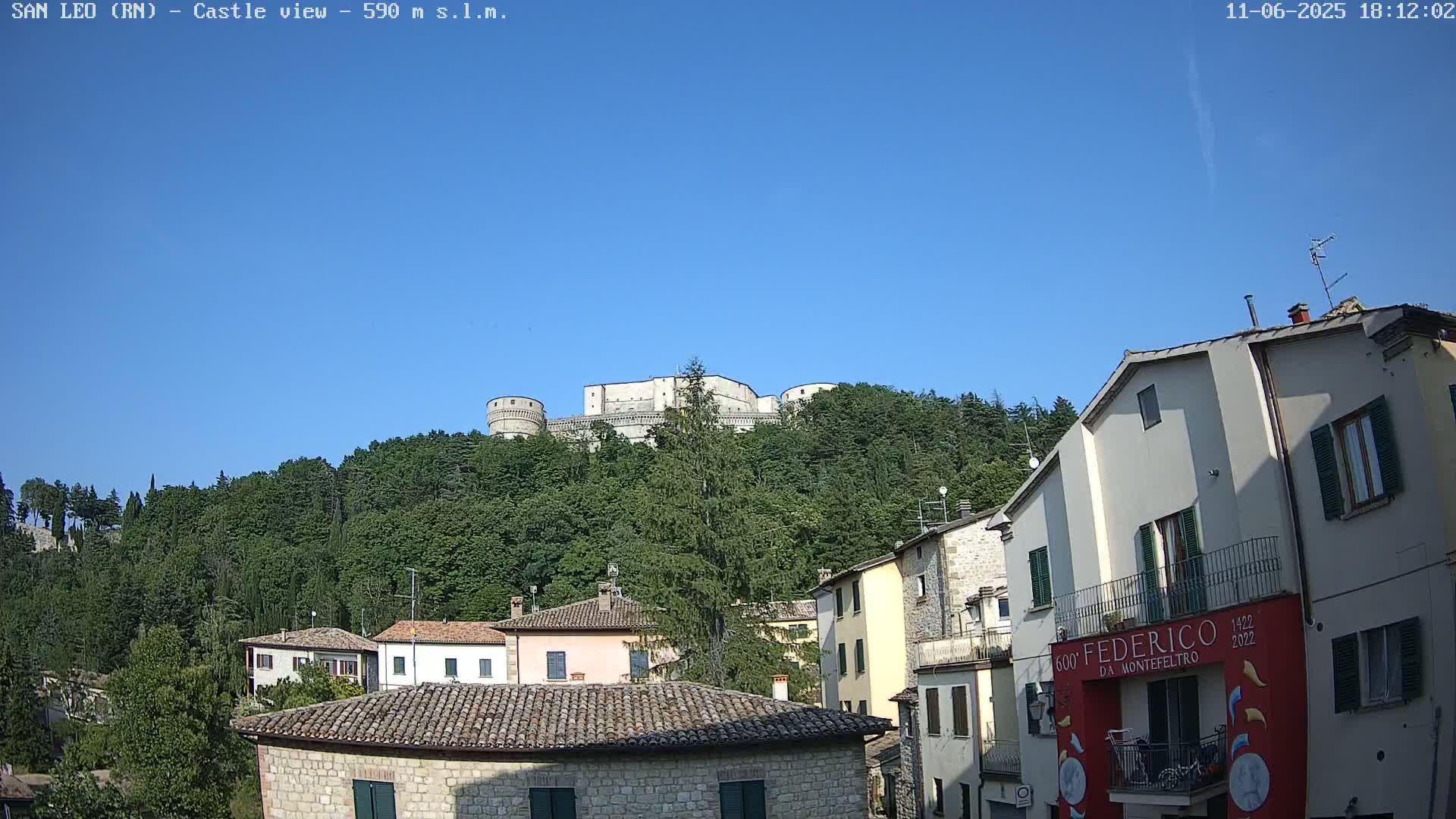 A stone castle sits atop a hill covered in lush green trees under a clear blue sky, overlooking a village of stone buildings.