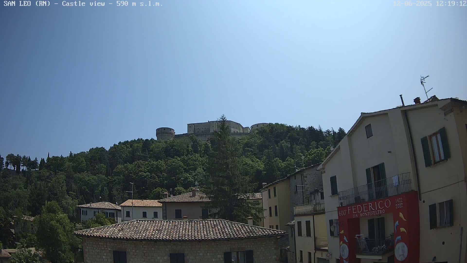 A stone castle sits atop a lush green hill overlooking a town of low stone buildings under a clear blue sky.