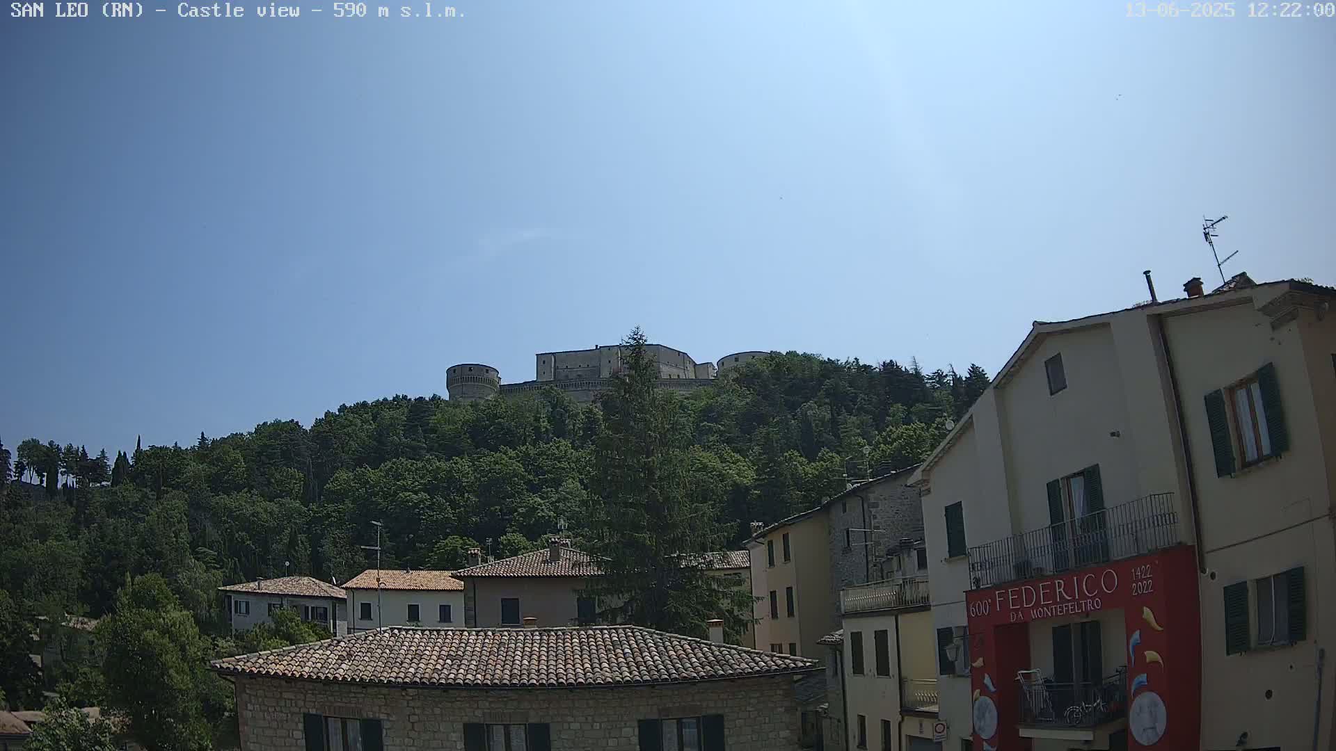 A stone castle sits atop a tree-covered hill overlooking a town of low stone buildings under a clear blue sky.