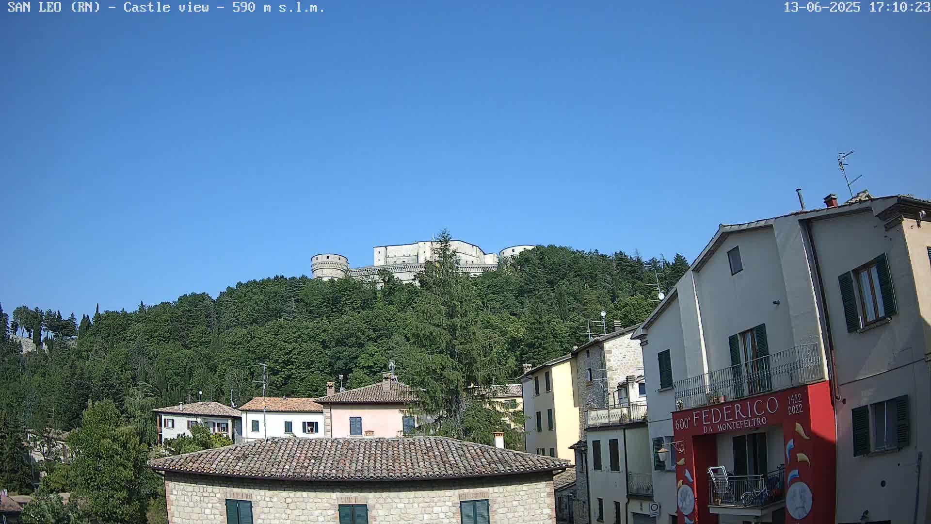 A stone castle sits atop a verdant hill overlooking a town of low stone buildings under a clear blue sky.