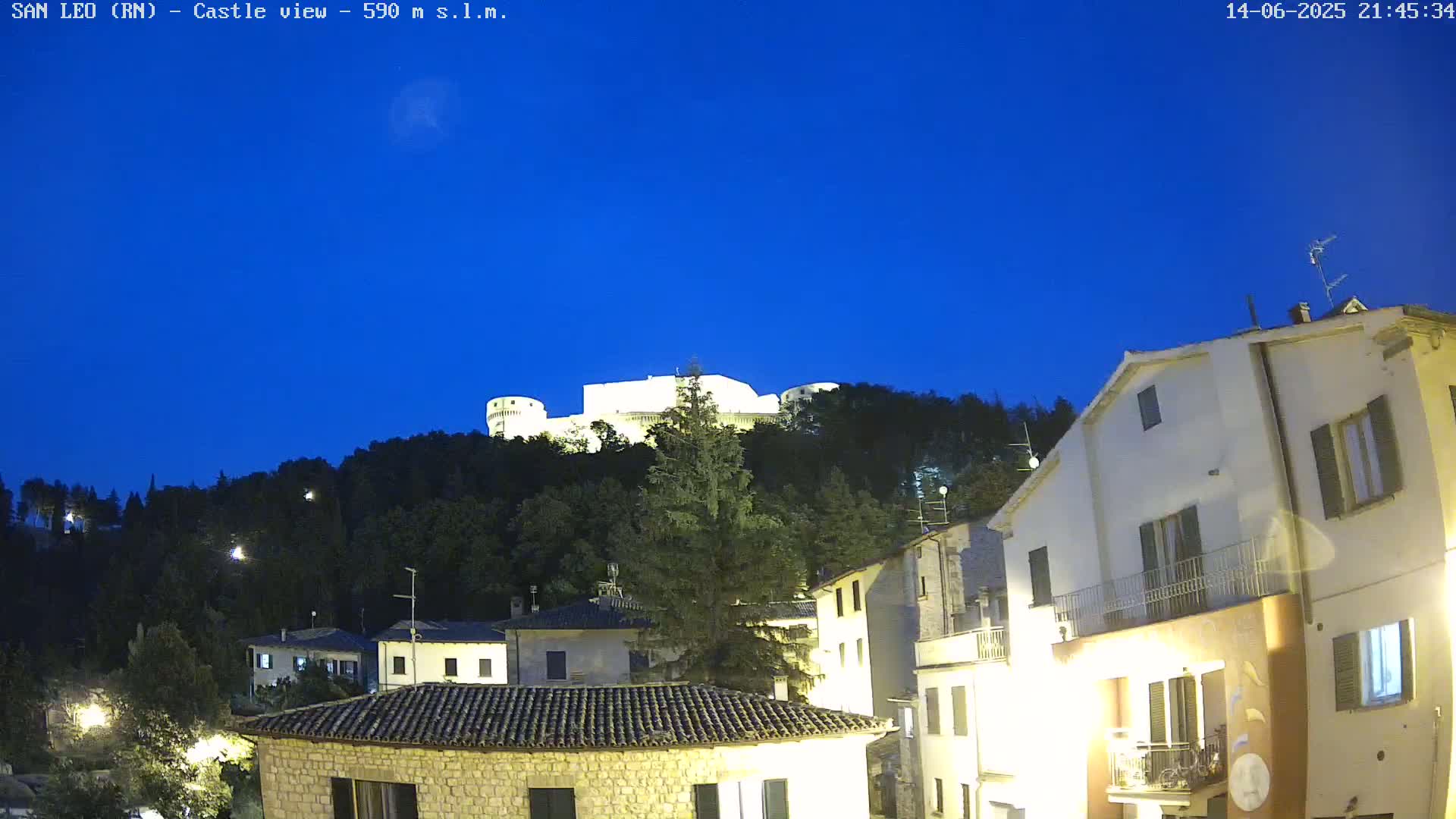 A stone castle sits atop a verdant hill overlooking a town of low stone buildings under a clear blue sky.
