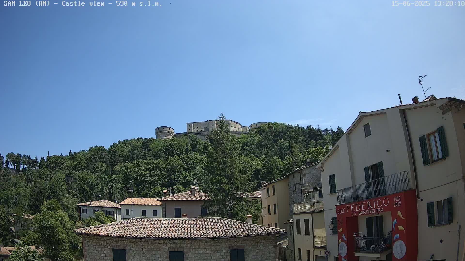 A stone castle sits atop a hill covered in lush green trees under a clear blue sky, overlooking a village of stone buildings with red tile roofs.