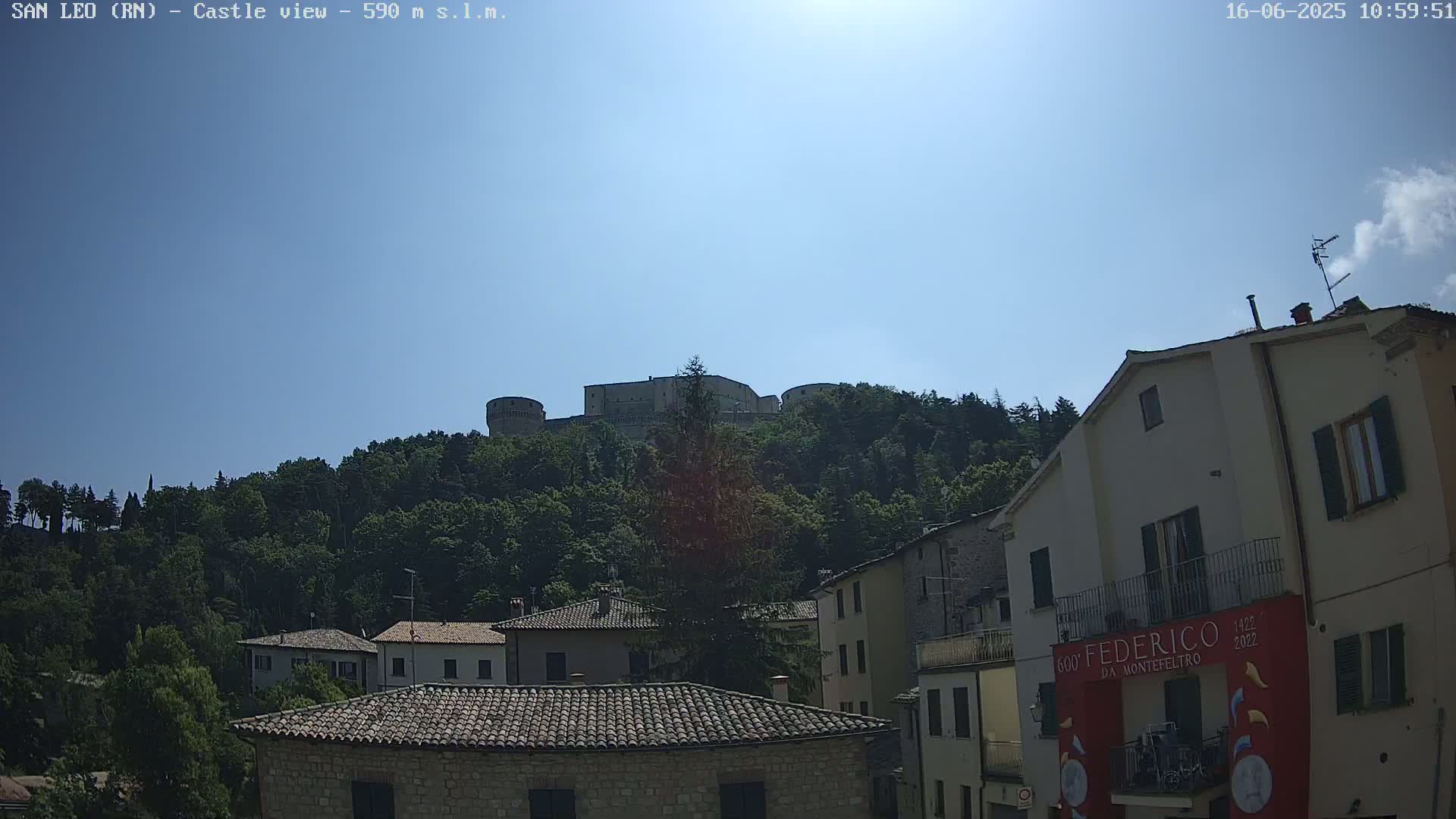 A stone castle sits atop a tree-covered hill overlooking a village of low buildings under a clear, sunny sky.