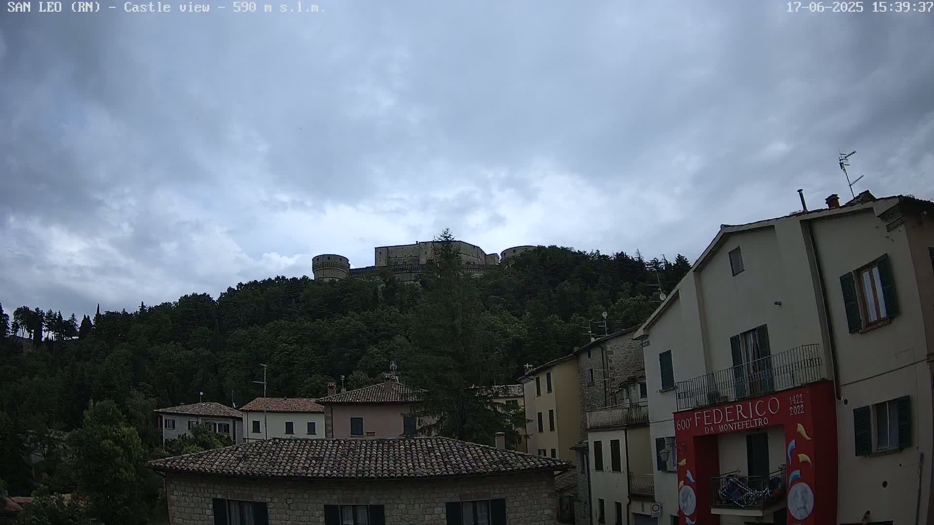 A stone castle sits atop a tree-covered hill overlooking a town of stone buildings under a mostly cloudy sky.