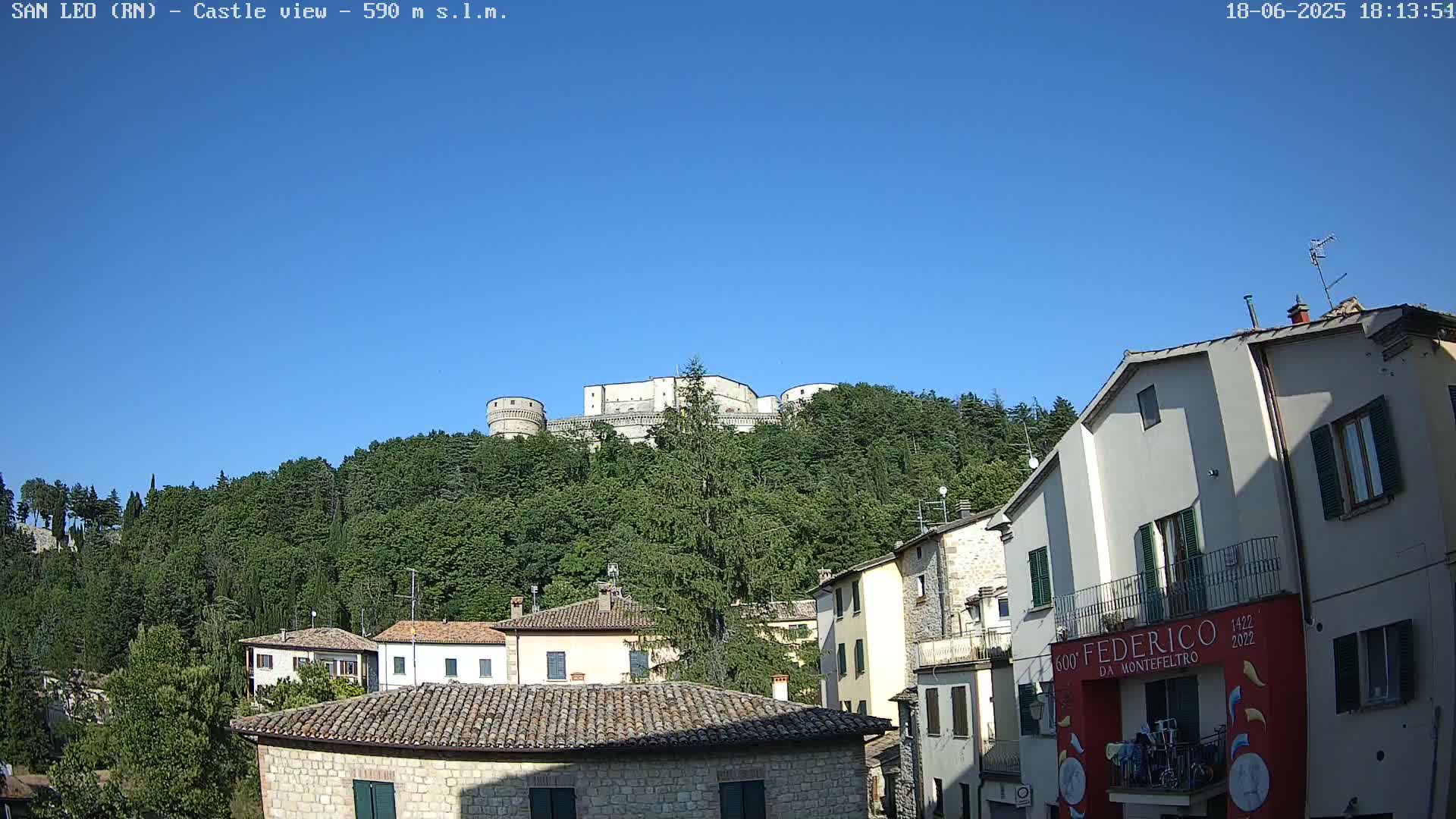 A stone castle sits atop a tree-covered hill overlooking a town of stone buildings under a clear blue sky.