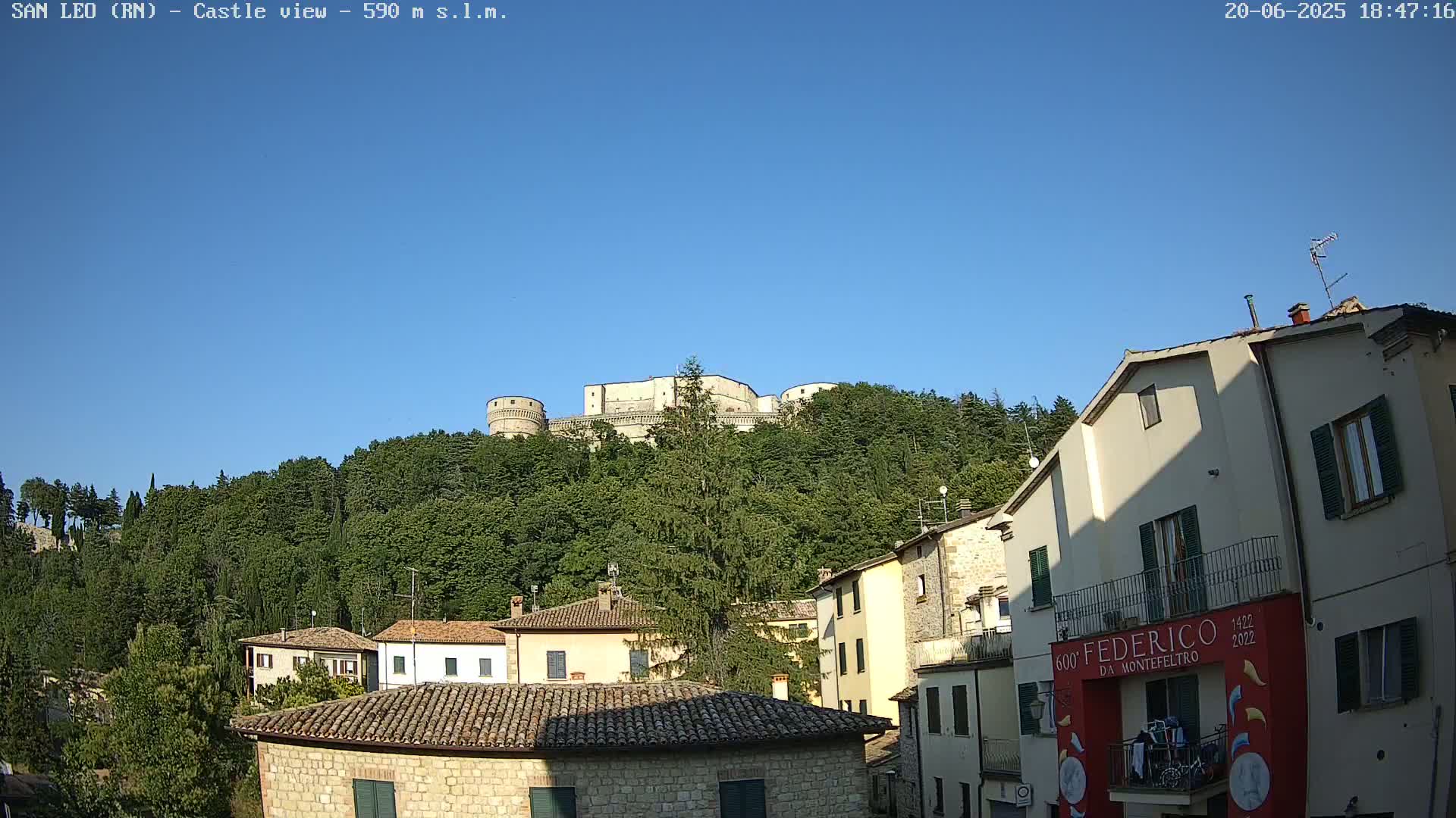 A stone castle sits atop a tree-covered hill overlooking a town of stone buildings under a clear blue sky.